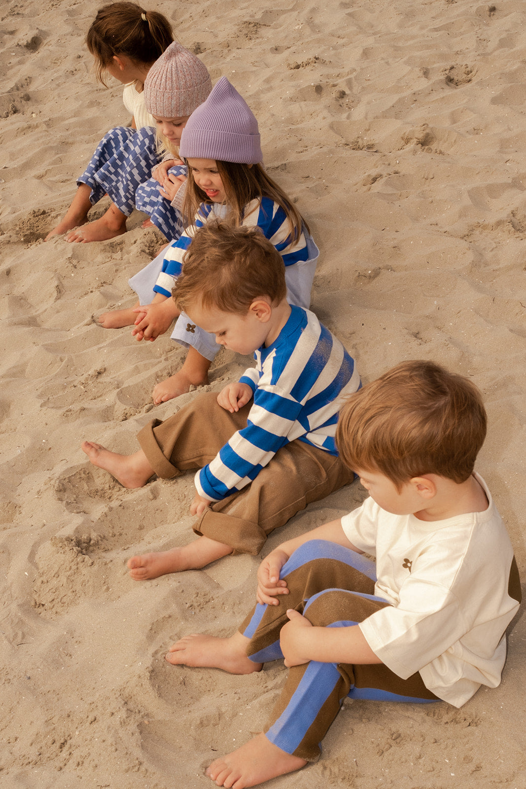 Five young children sit barefoot on the sandy beach, wearing GROWNβs Stripe Long Sleeve Tee in cozy earth-toned yarn dyed stripes and lightweight jersey, enjoying a calm moment as they look down or play with the sand together.