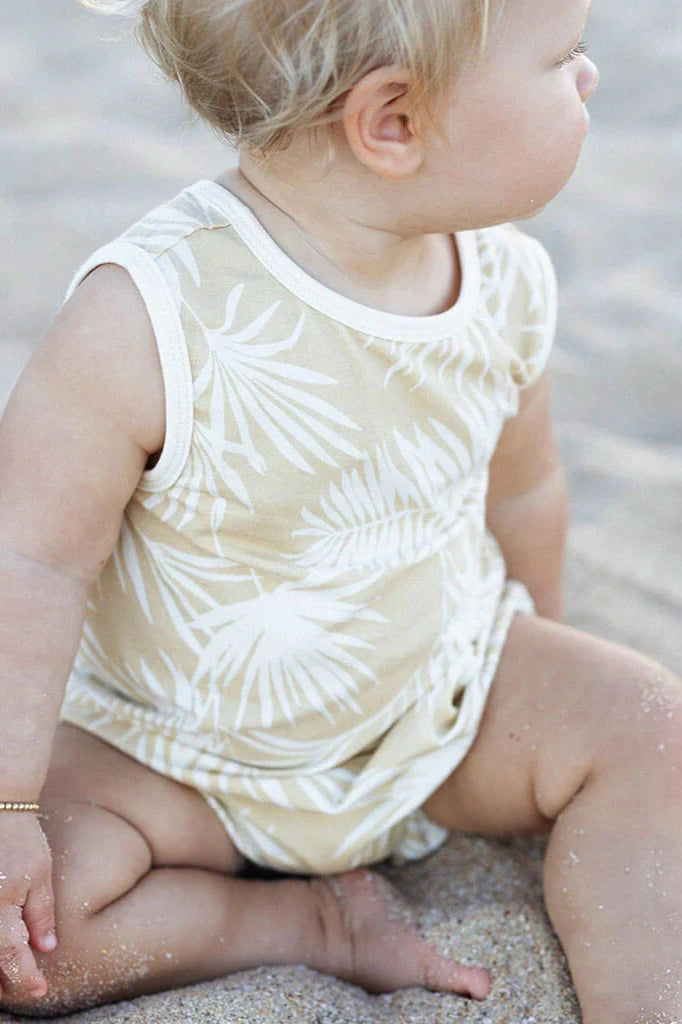 A toddler with blond hair sits on the sandy ground at the beach, dressed in a Hula Playsuit by BAM LOVES BOO. Their skin is sprinkled with sand as they gaze away from the camera, perfectly capturing a moment of seaside innocence.