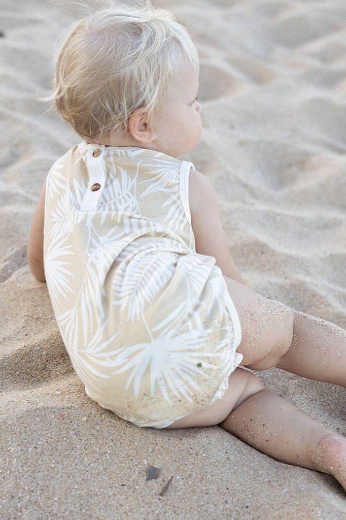 A toddler with blonde hair sits on the sand at a beach, wearing the Hula Playsuit by BAM LOVES BOO, featuring a beige and white vintage palm print. The super soft 2-way stretch fabric and eco-friendly dyes make it perfect for play. The child gazes away from the camera as sand lightly covers their legs.