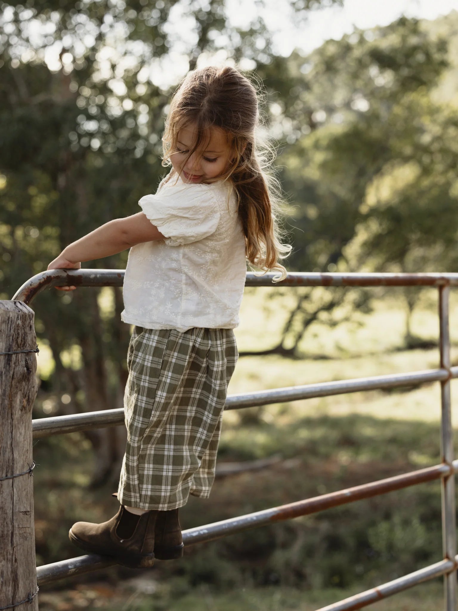 A young child with long hair, wearing a white shirt, plaid skirt, and brown boots, stands on a metal fence in a rural outdoor setting. With trees and greenery blurred in the background creating a serene, natural environment, the child's outfit could easily include the whimsical unisex Rio Pant Olive by VALENCIA BYRON BAY, perfect for any adventure.