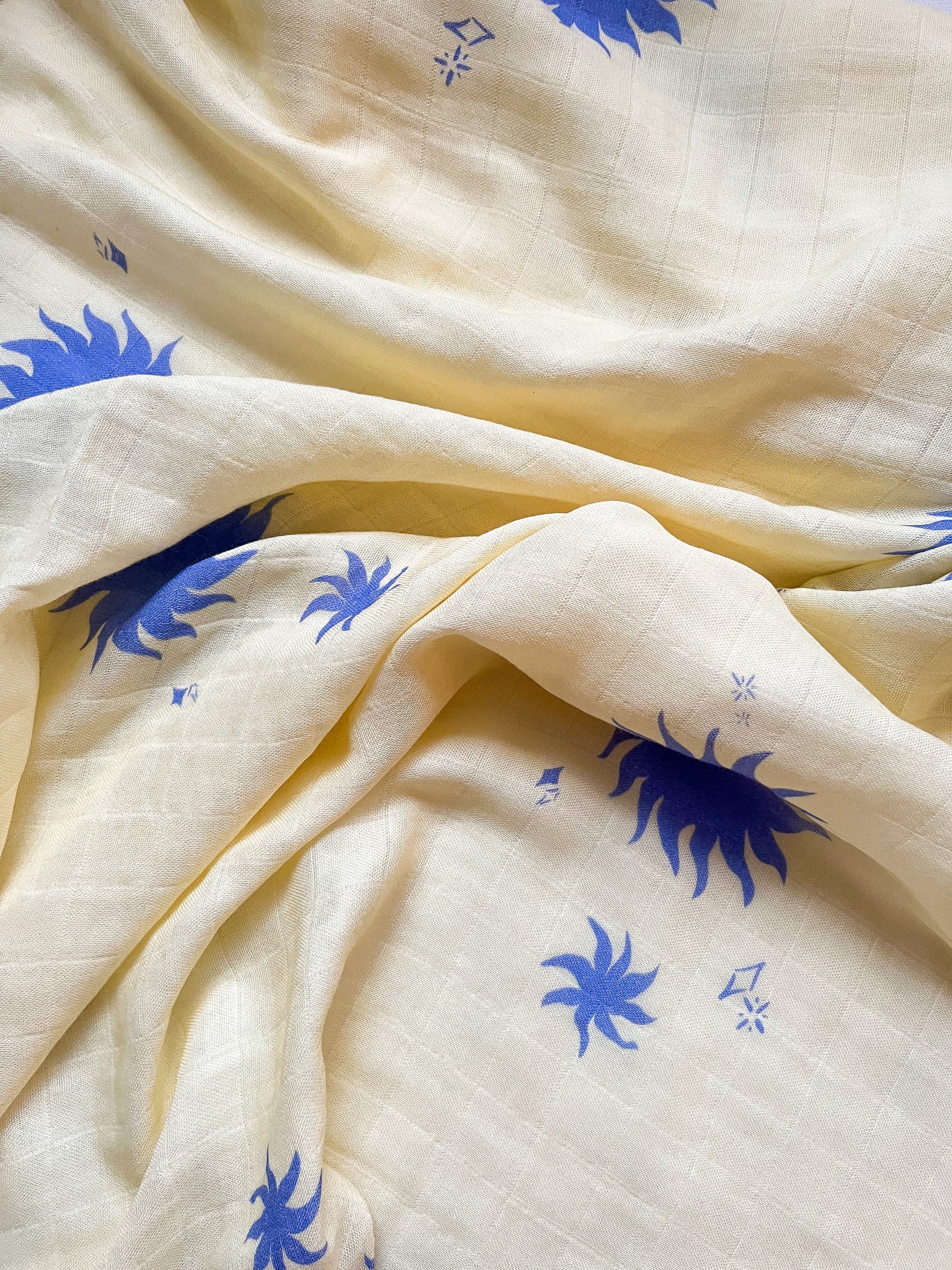The MILKY DESIGNS Sol Wrap is a soft, cream-colored bamboo cotton baby blanket with a subtle grid texture, exclusive blue sun motif prints, and scattered geometric shapes for breathable comfort.