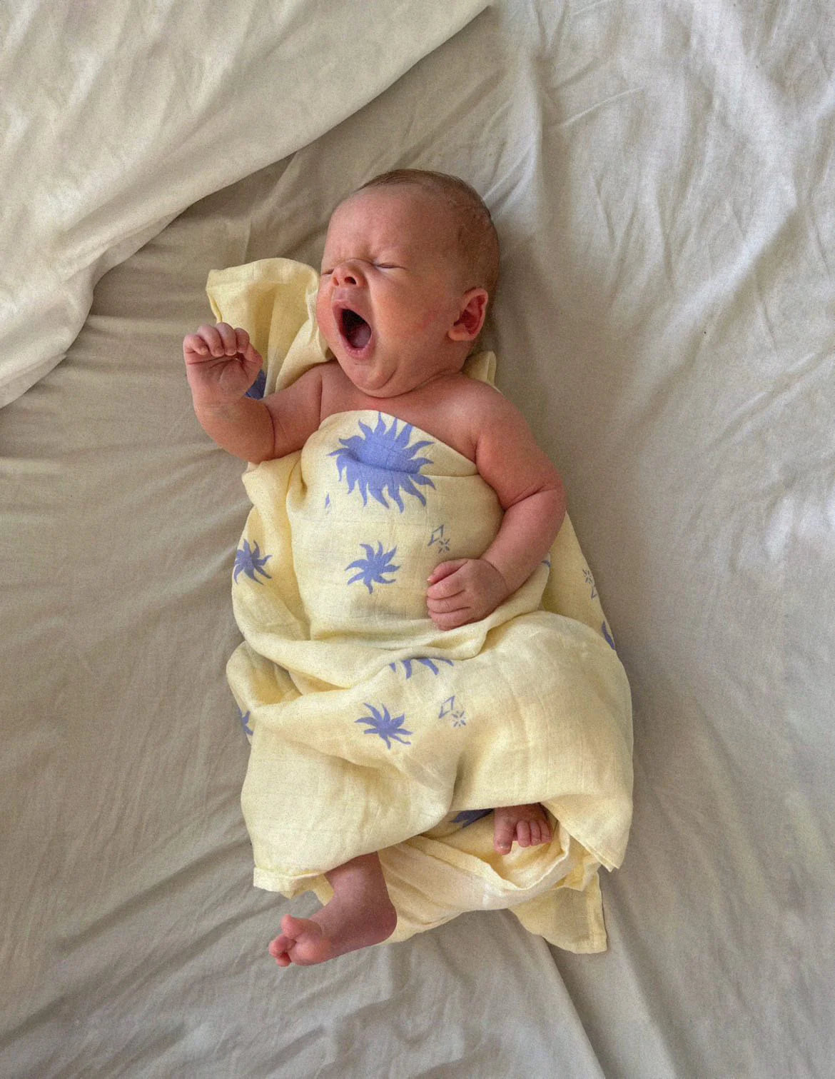 A newborn swaddled in the Milky Designs - Sol Wrap by MILKY DESIGNS, featuring exclusive yellow fabric with blue sun prints, lies on a white bed yawning with eyes closed.