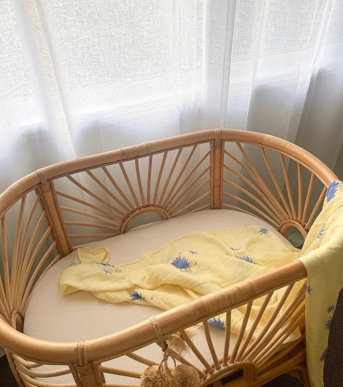 A rattan baby bassinet sits by a window with sheer white curtains. A Milky Designs - Sol Wrap from MILKY DESIGNS, featuring soft yellow and blue floral prints, is draped inside as natural light softly fills the space.