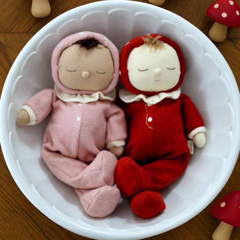 Two OLLI ELLA Dozy Dinkum dolls, each with closed eyes—one in pink, one in red—rest side by side in a white bowl. Olli Ella - Dozy Dinkum Toadstool Pip wooden mushrooms are scattered around on the wooden surface.
