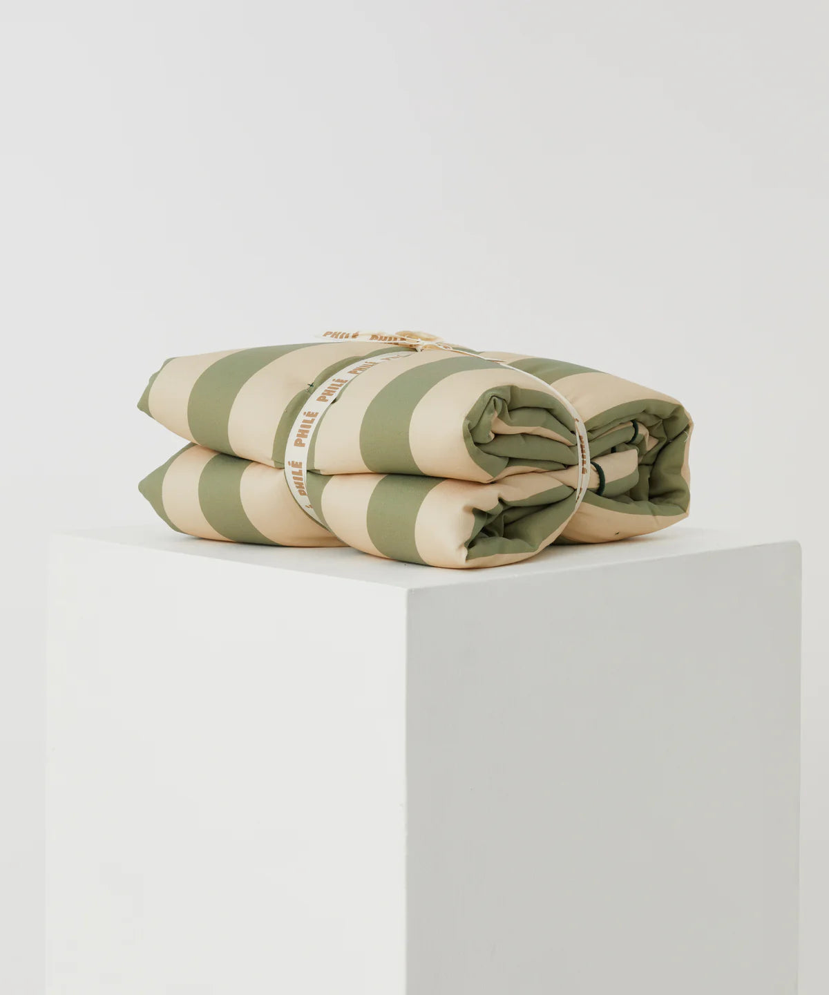 The Philé - Striped Play Mat Green by PHILÉ, made with eco-friendly OEKO-TEX cotton and wide green and beige stripes, is folded, tied with a white ribbon, and displayed on a white pedestal against a light background.