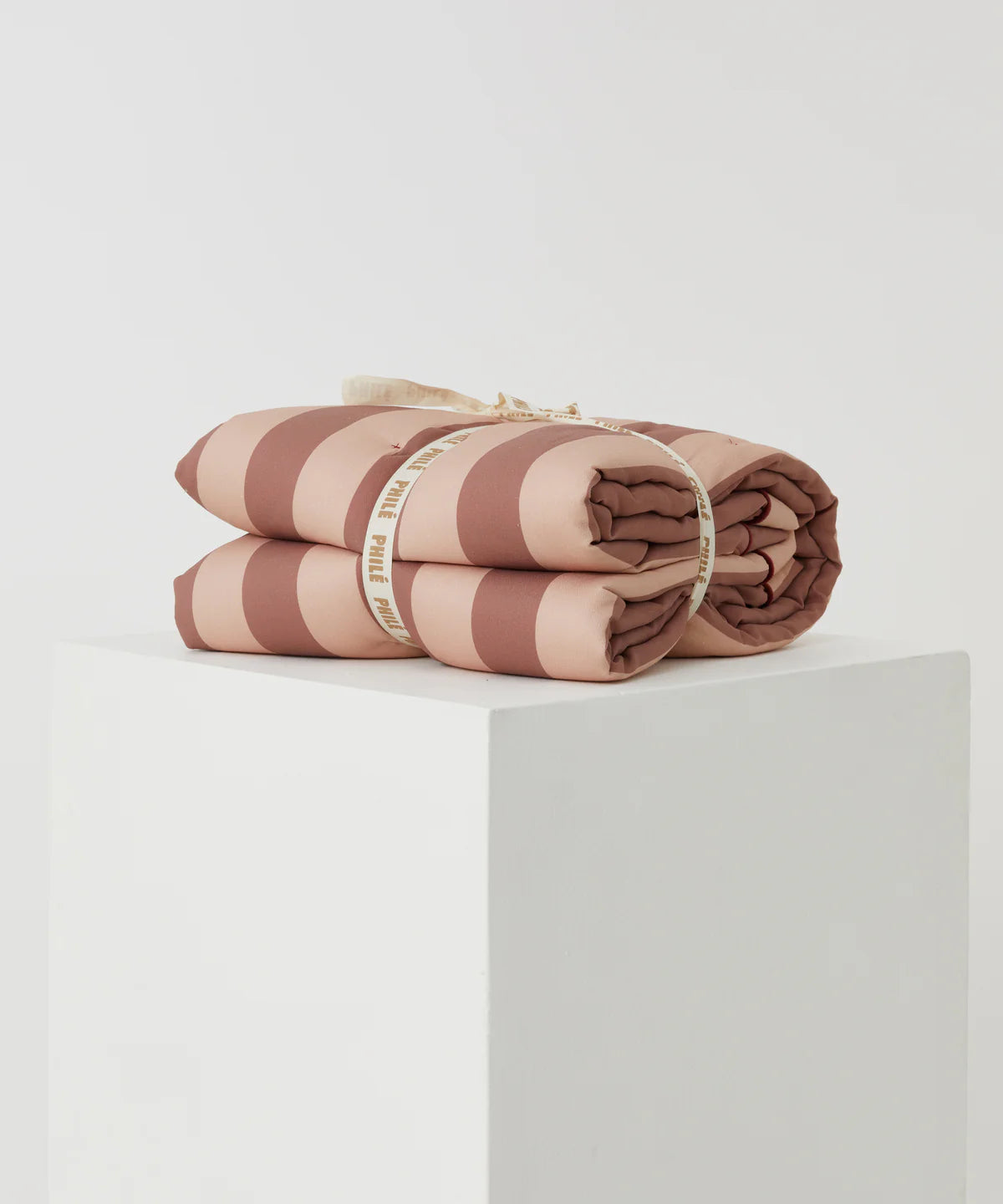 The Philé - Striped Play Mat Pink by PHILÉ, crafted from eco-friendly OEKO-TEX Cotton in pink and brown stripes and tied with a white ribbon, sits folded on a white pedestal against a plain light background.