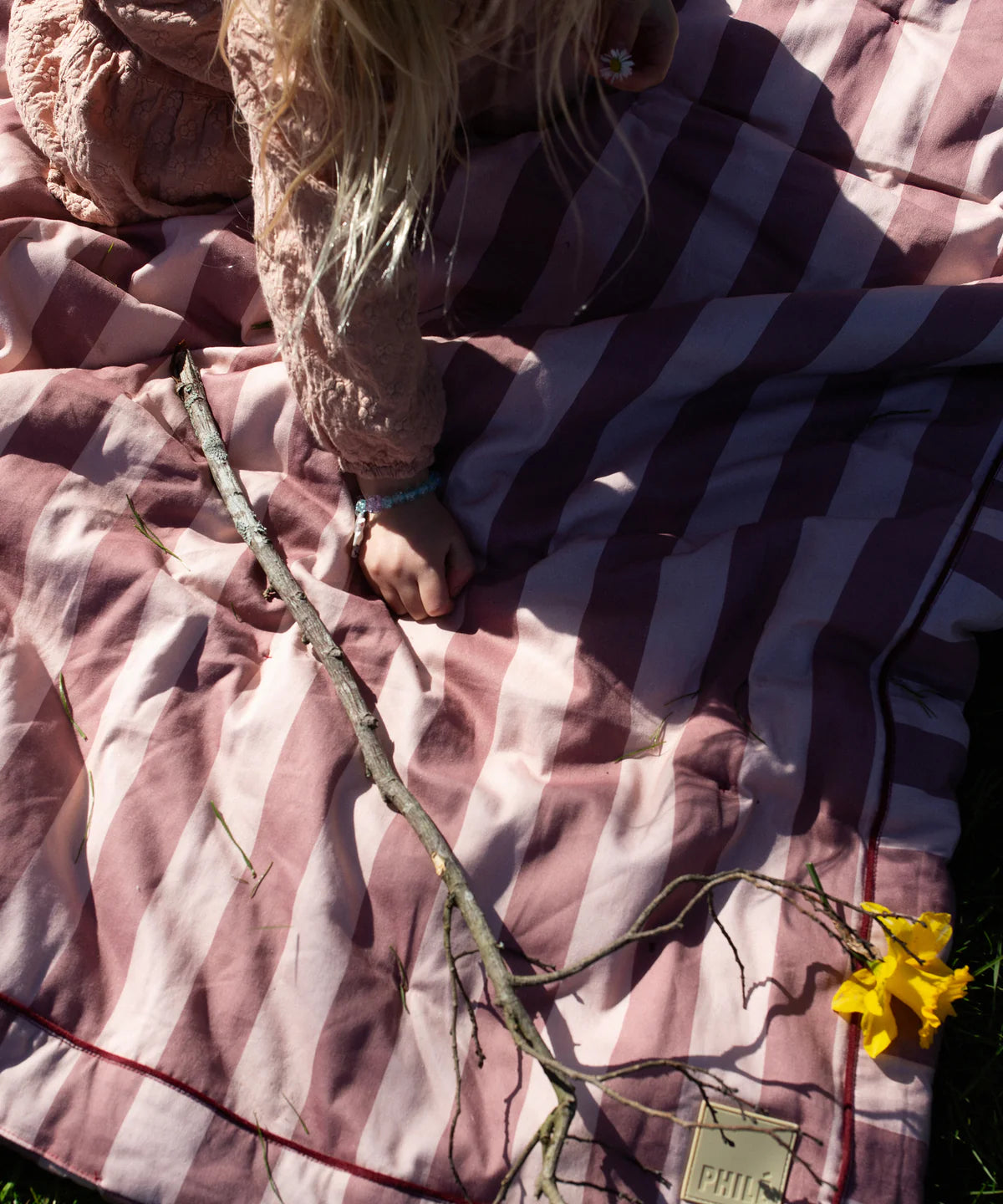 A child with long blond hair sits outdoors on the PHILÉ Philé - Striped Play Mat Pink, reaching for a stick and a yellow flower as sunlight casts soft shadows.