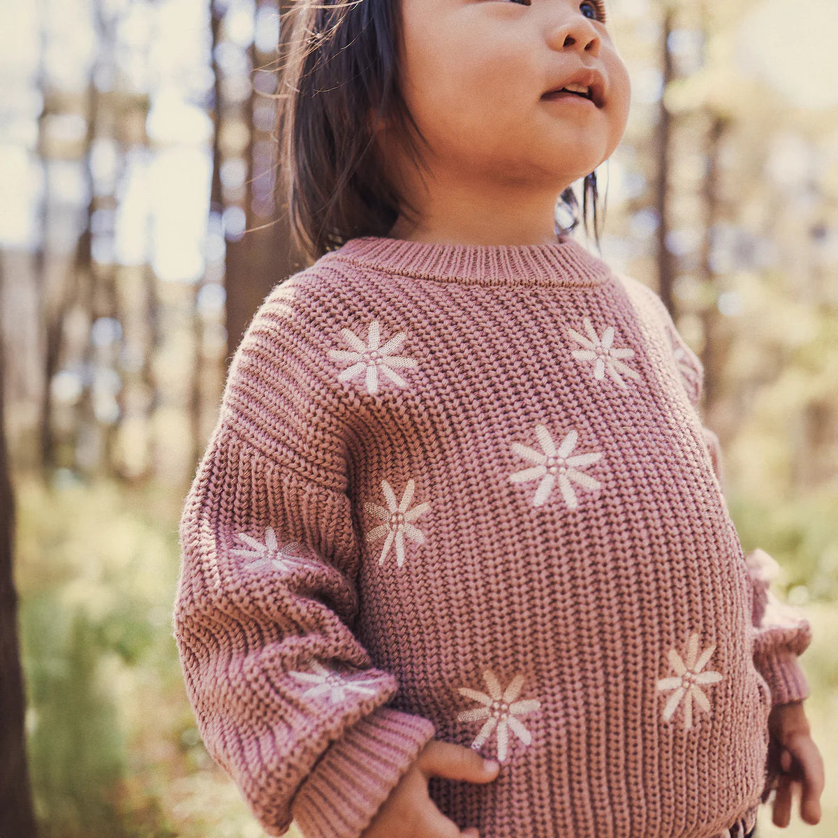 A young child wears the KYND BABY Kynd Baby - Embroidered Knit Jumper Wood Rose, featuring delicate white flower embroidery, while standing outdoors in a sunlit forest and smiling.