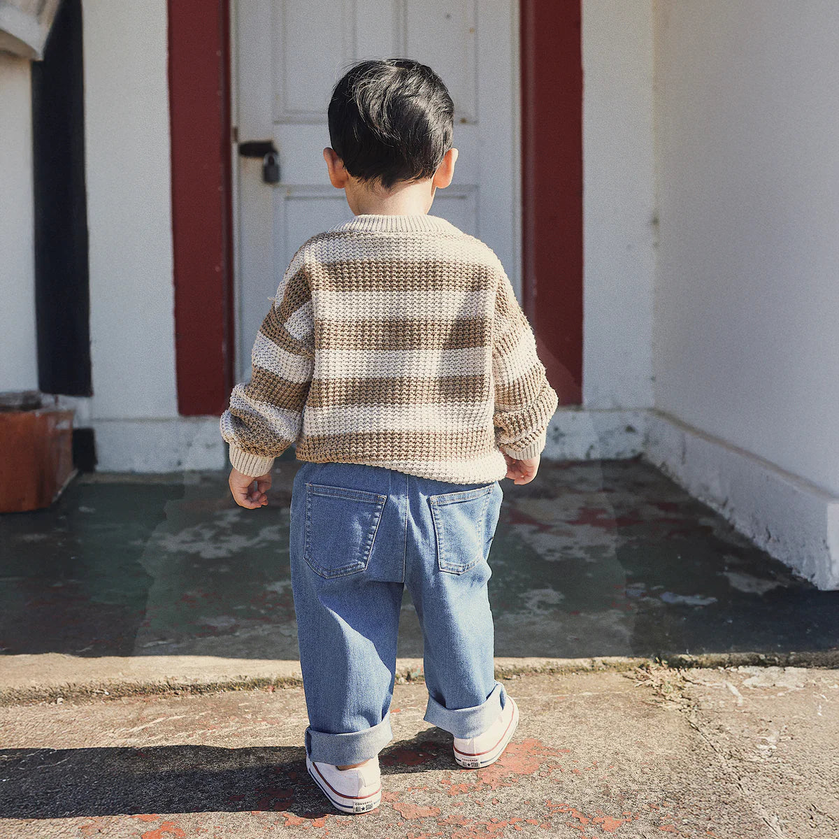 A toddler with short dark hair, seen from behind in sunlight, wears the KYND BABY Organic Textured Knit Jumper Caramel Stripe, blue jeans, and sneakers while standing by a white door outside a house.