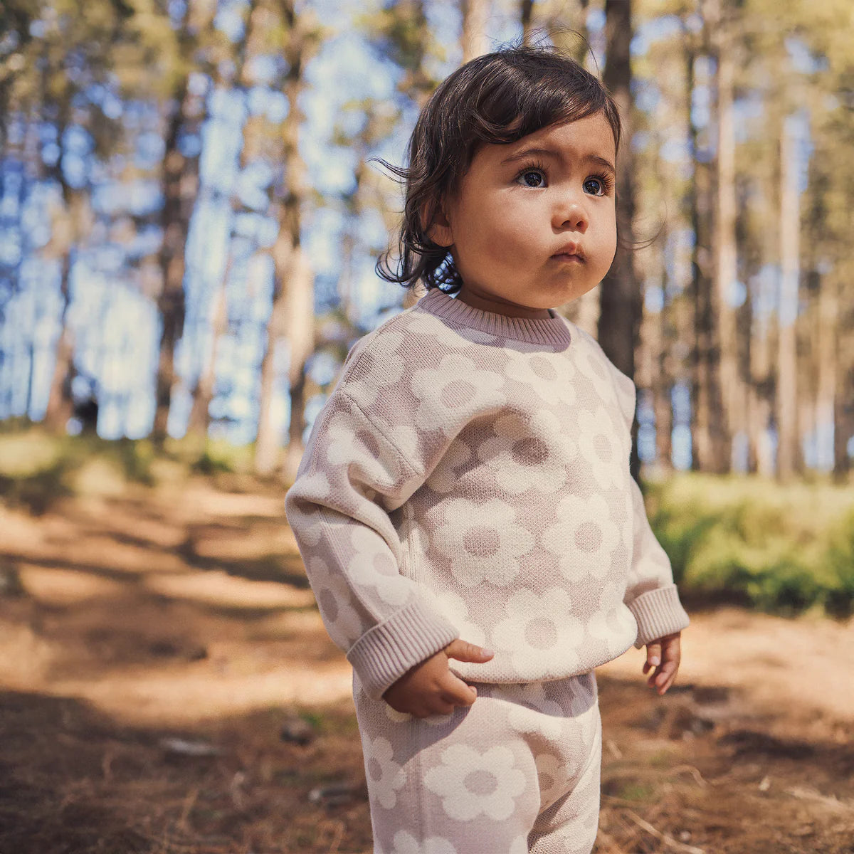 A young child wears the KYND BABY Kynd Baby - Knit Jumper Neutral Flower, featuring white floral patterns, while standing in a sunlit forest with tall trees and dappled light in the background.