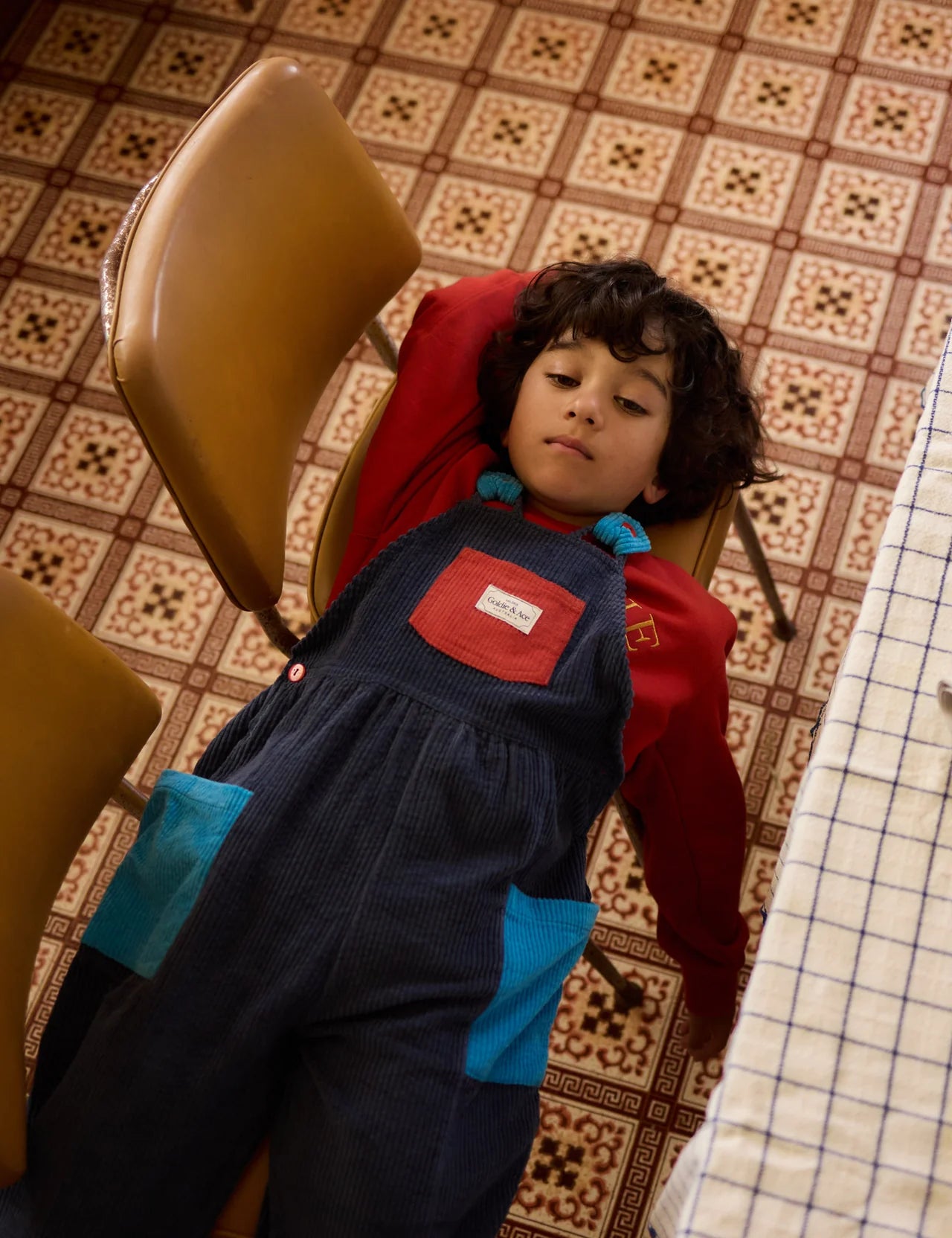 A young boy with curly hair relaxes on a patterned floor, wearing GOLDIE + ACE's Goldie + Ace - Sammy Corduroy Colour Block Overalls—a stylish piece with adjustable straps and oversized pockets—paired with a red long-sleeve shirt.