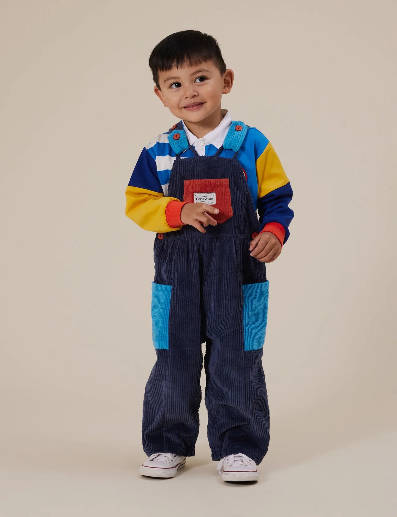 A young child smiles, wearing GOLDIE + ACE’s Sammy Corduroy Colour Block Overalls with adjustable straps and oversized pockets, styled with a white collared shirt and a vibrant striped sweatshirt in blue, yellow, and red.