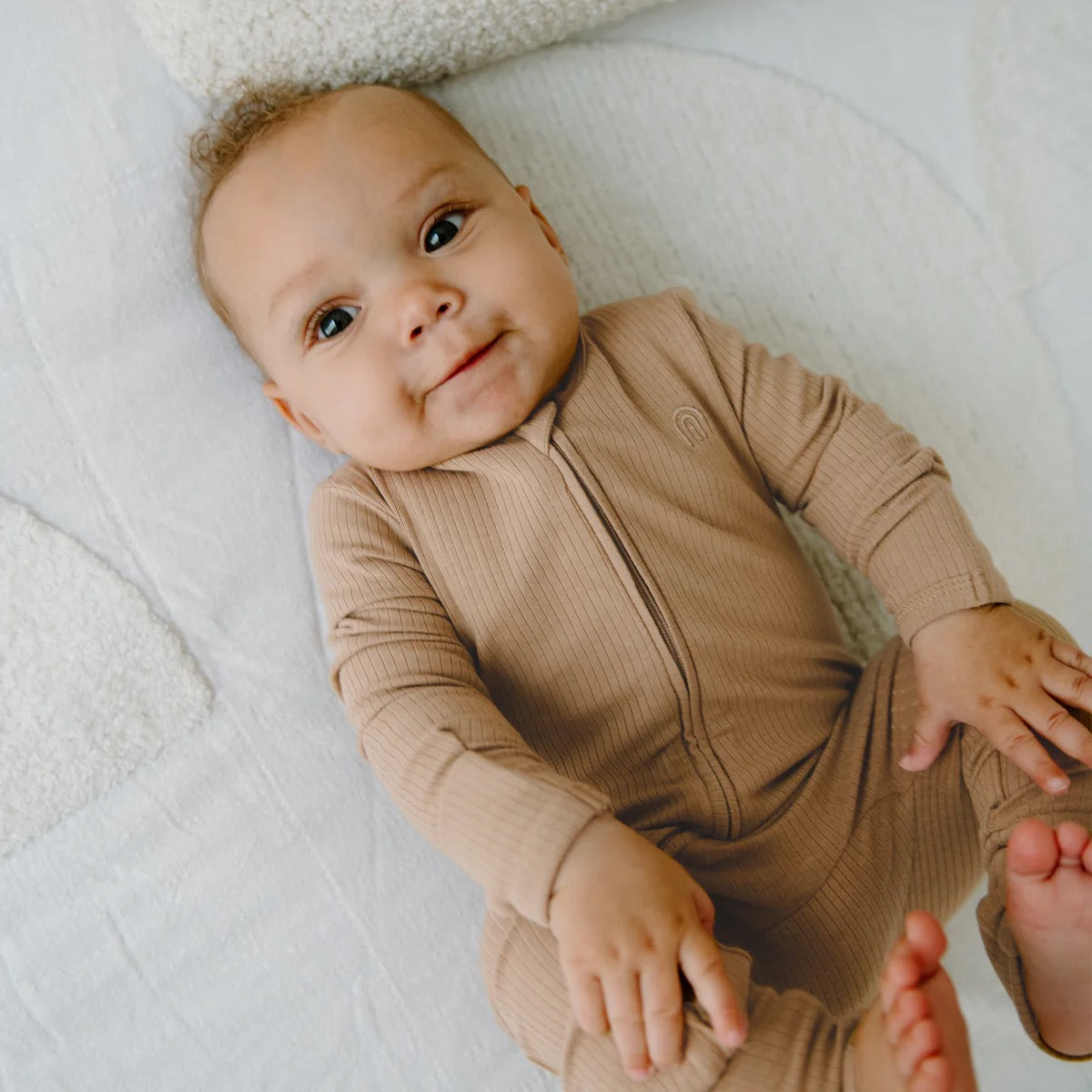 A smiling baby with curly hair wears the KYND BABY Rib Day or Night Onesie Mocha, featuring a 2-way zipper, while lying on a white textured blanket and gazing gently upward.