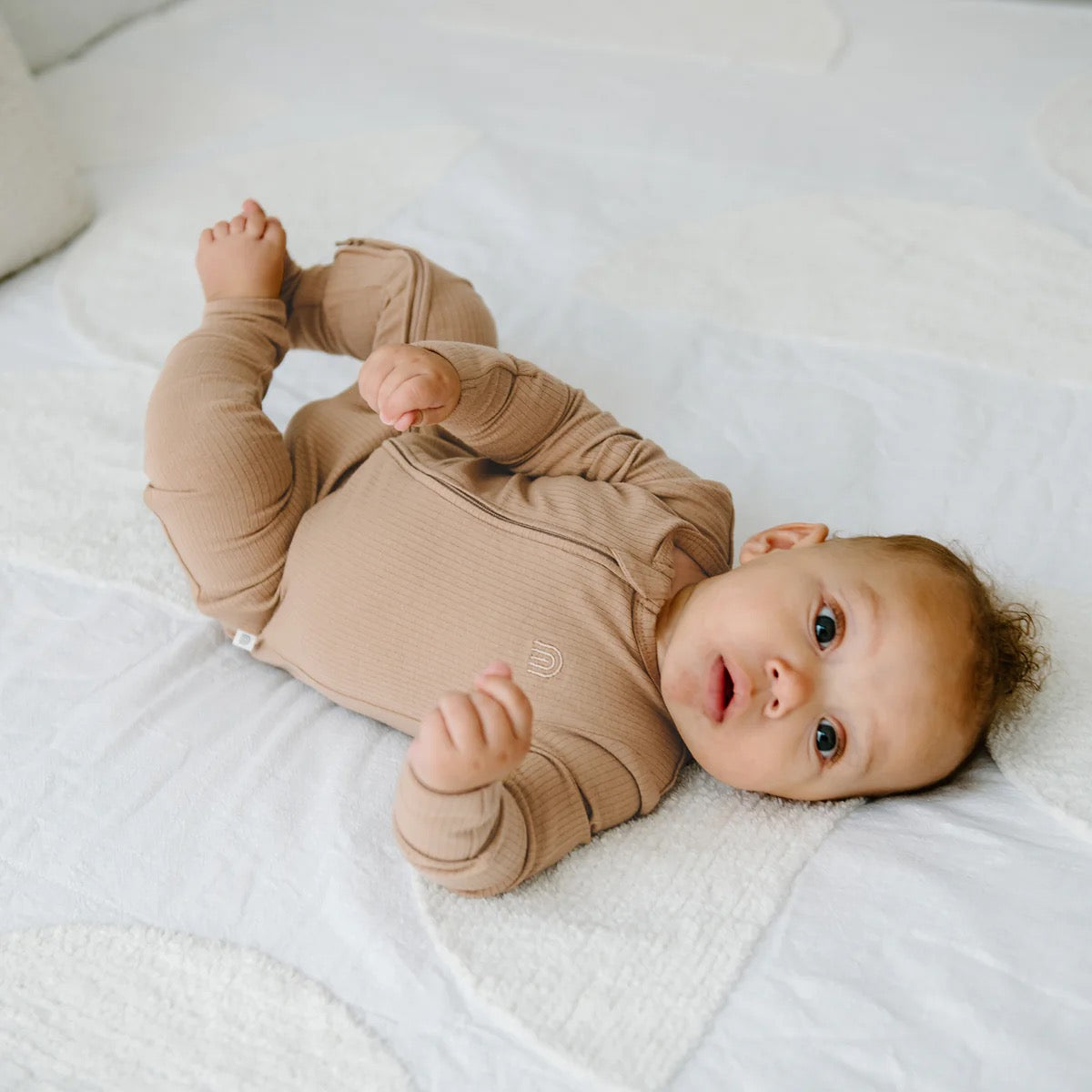 A baby in a Kynd Baby - Rib Day or Night Onesie Mocha by KYND BABY with a 2-way zipper lies on a white textured blanket, gazing upward with wide eyes and slightly raised arms.