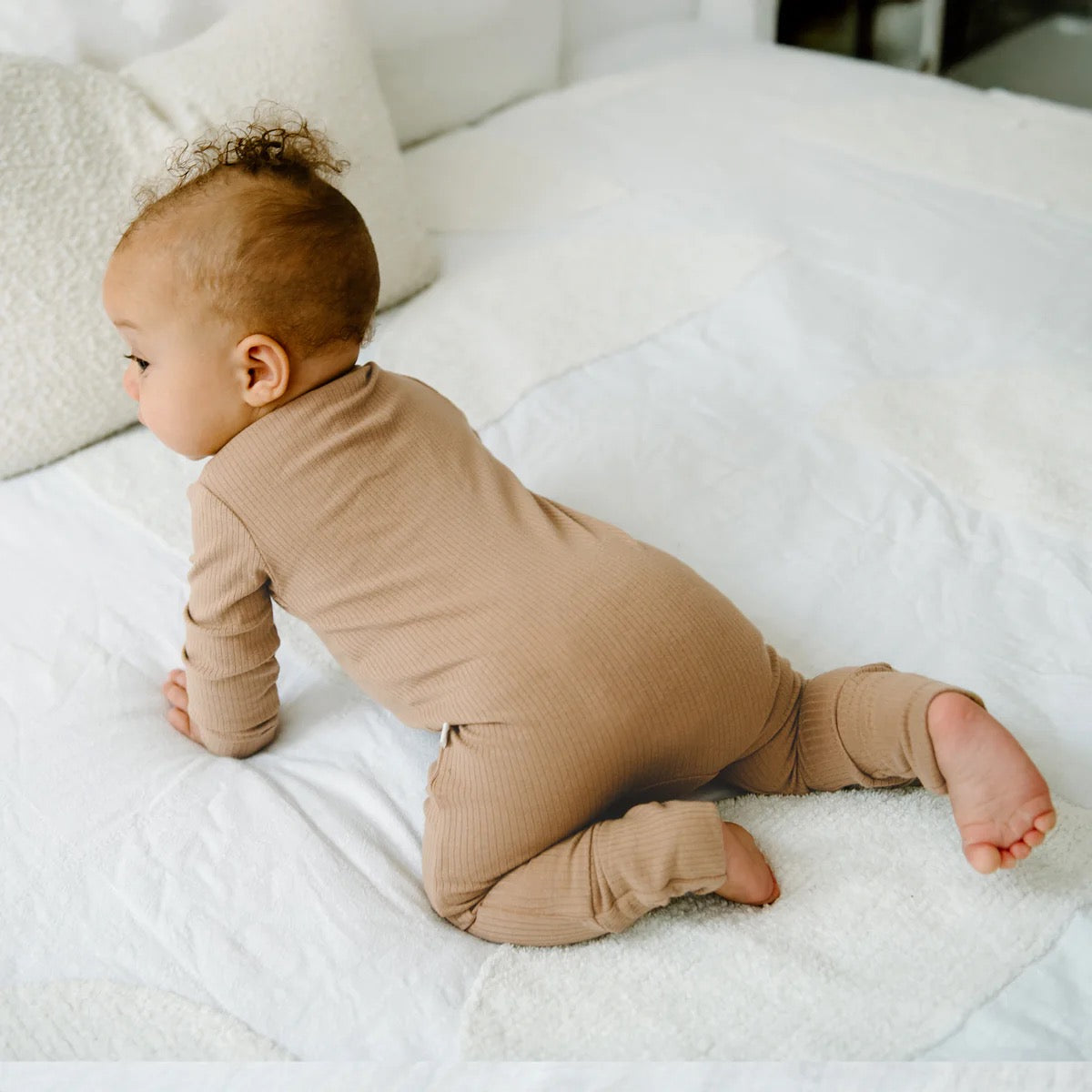 A baby in the KYND BABY Rib Day or Night Onesie Mocha crawls on a white bed, surrounded by soft pillows and blankets that enhance the cozy atmosphere.