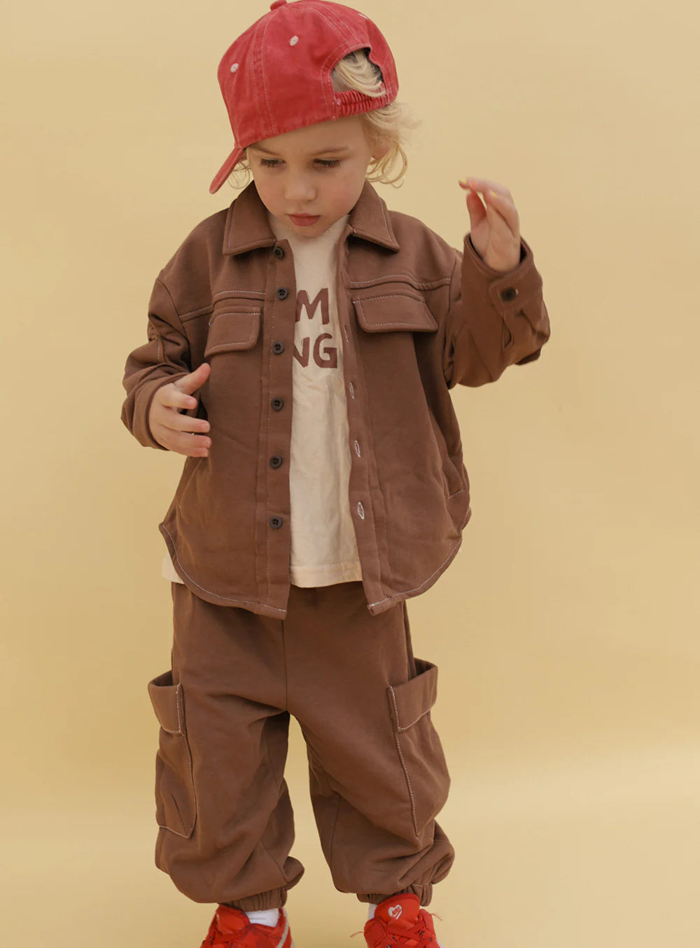 A young child wears Small Swim Club - Choccy Cargo Trackies by SMALL SWIM CLUB, paired with a cream t-shirt, red sideways cap, and red shoes. The child stands against a beige background, looking down with one hand raised.