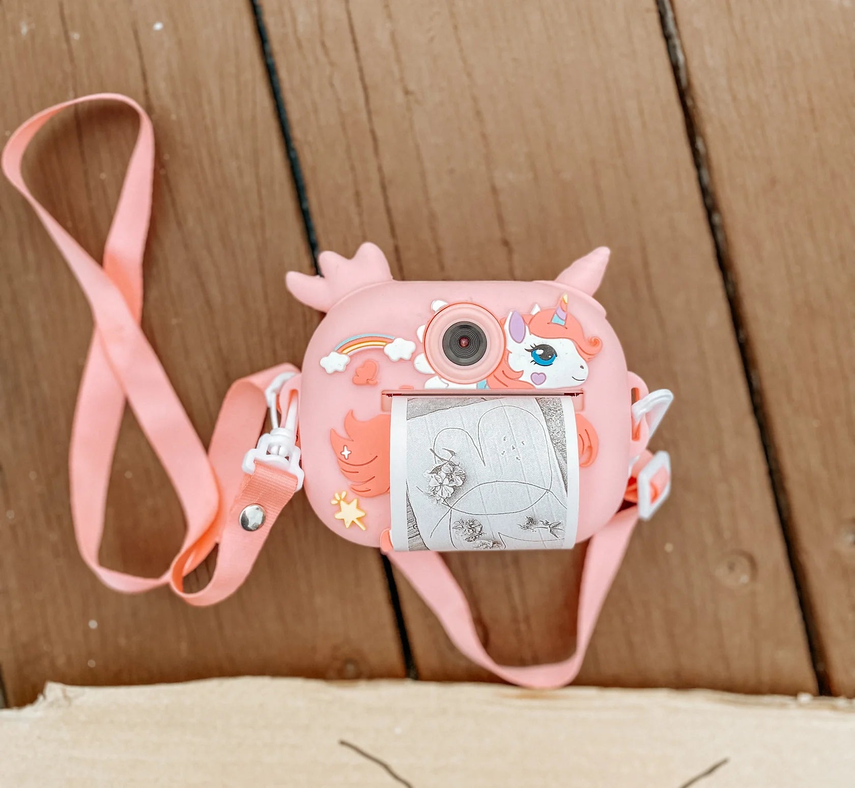 The OH MY FRANKIE Oh My Snap Instant Print Camera Pink features a unicorn design, prints black-and-white photos, and comes with a pink strap for easy carrying. It rests on a wooden floor with stars and a rainbow for decoration.