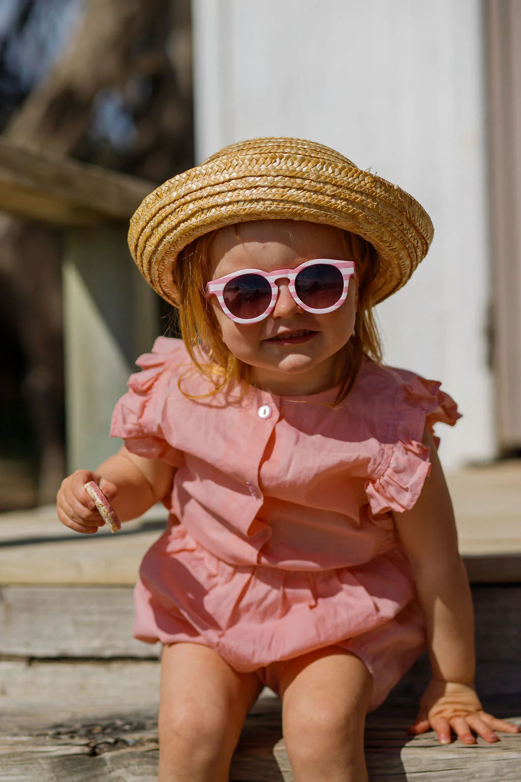 A young child sits on wooden steps wearing a pink outfit, a straw hat, and FRANKIE RAY's Baby Pixie Sunglasses Pink Stripe with impact-resistant lenses. The child holds a small object in one hand and enjoys the outdoors with blurred trees in the background.