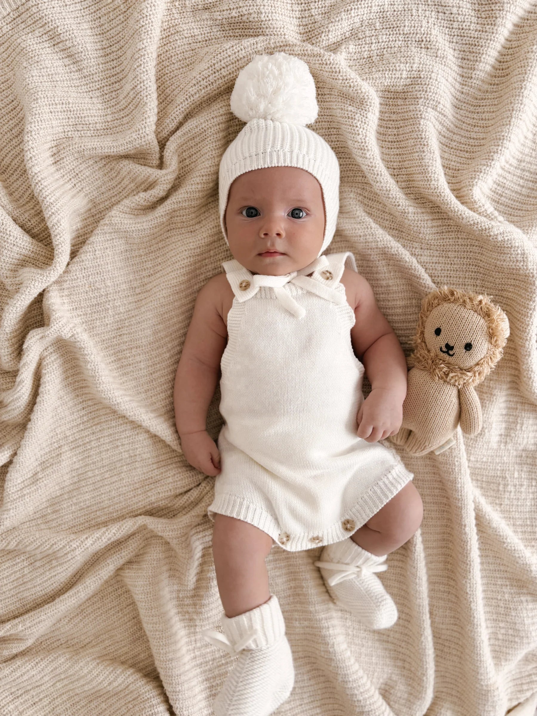 A baby in ZIGGY LOU’s Milk Booties, a cream knit romper, and pom-pom hat lies on a textured beige blanket beside a stuffed lion toy.