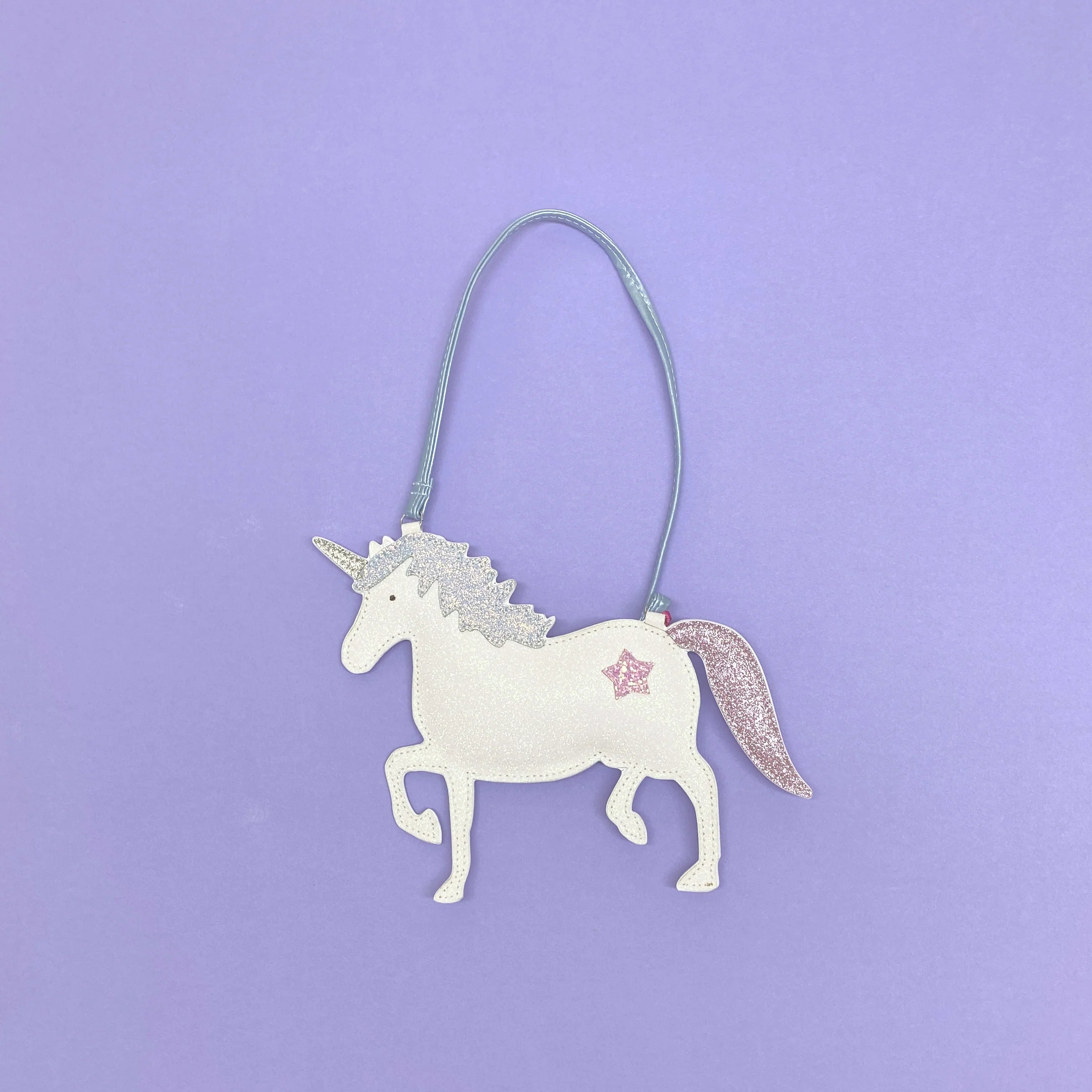 The Rockahula - Unicorn Glitter Bag by ROCKAHULA features a sparkly white unicorn with a pink tail, silver mane, and a pink star on its side, hanging from a metallic blue strap against a pastel purple background.