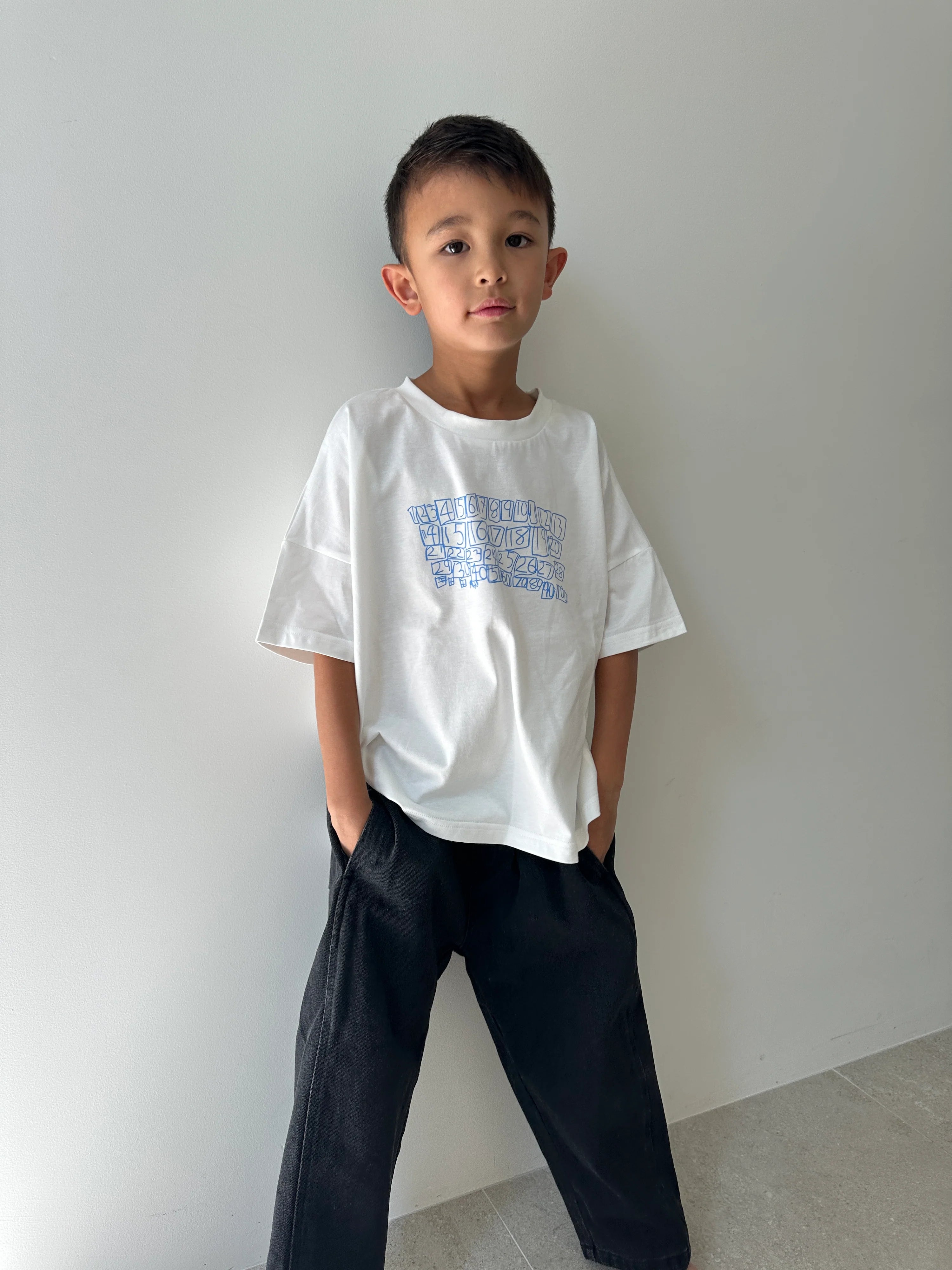 A young boy with a neutral expression stands against a light-colored wall, wearing the TINY TROVE Parker Numbers Relaxed Tee in white/blue and black pants, his hands in his pockets. The tee is made from 100% cotton jersey.