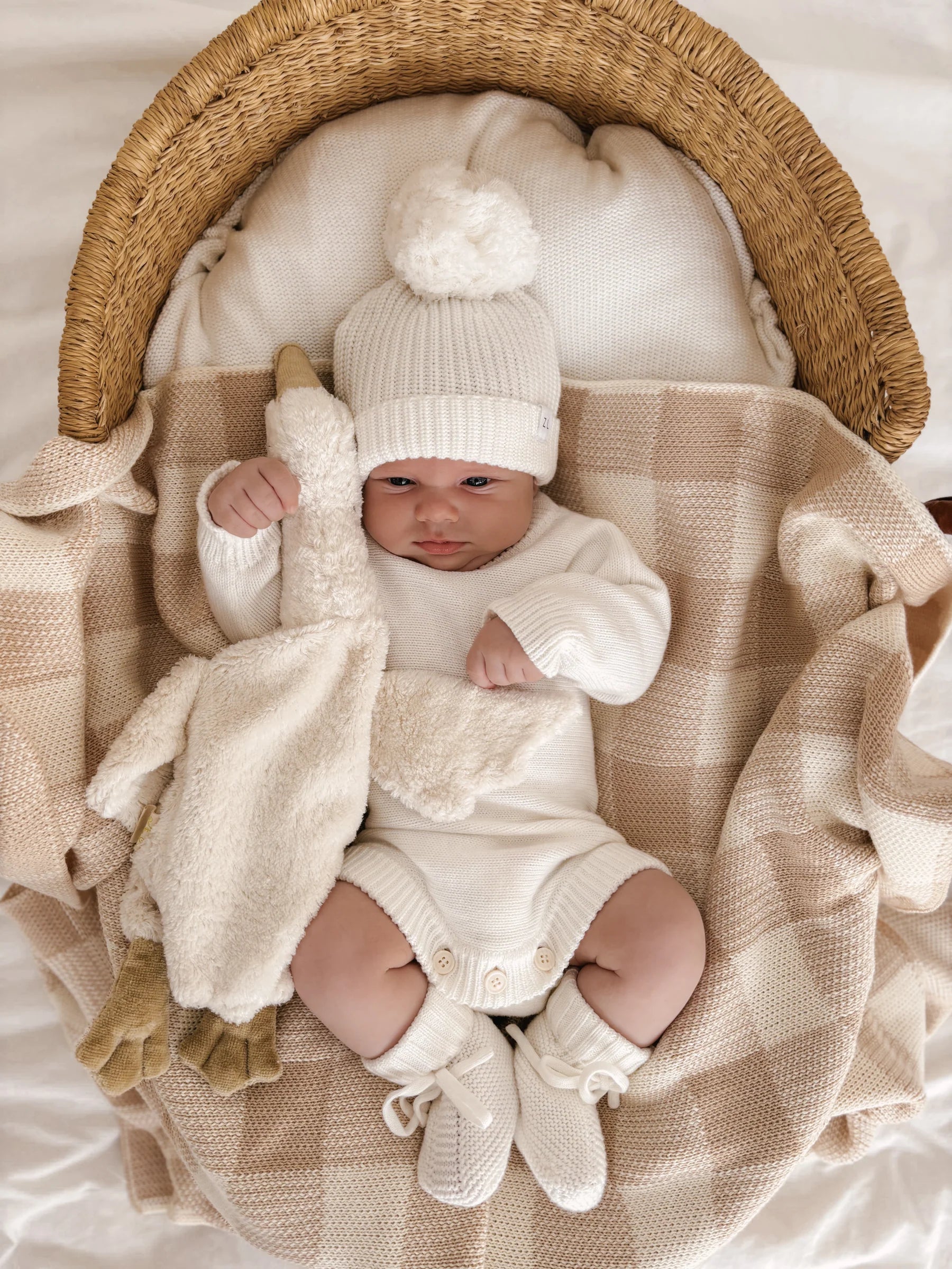 A baby in cozy knitwear and hat lies in a basket lined with the ZIGGY LOU Ziggy Lou - Blanket Gingham, a soft, machine washable cotton blanket—perfect for newborn safety and comfort.