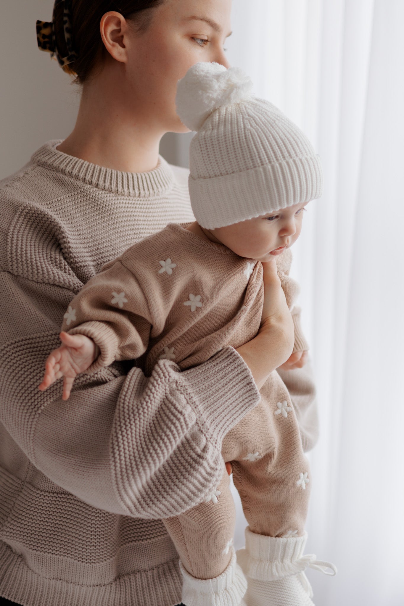 A woman in a beige sweater holds a baby wearing the ZIGGY LOU - Winter Playsuit Aurora (Size NB) and a white knit hat, standing by a bright window with soft natural light.