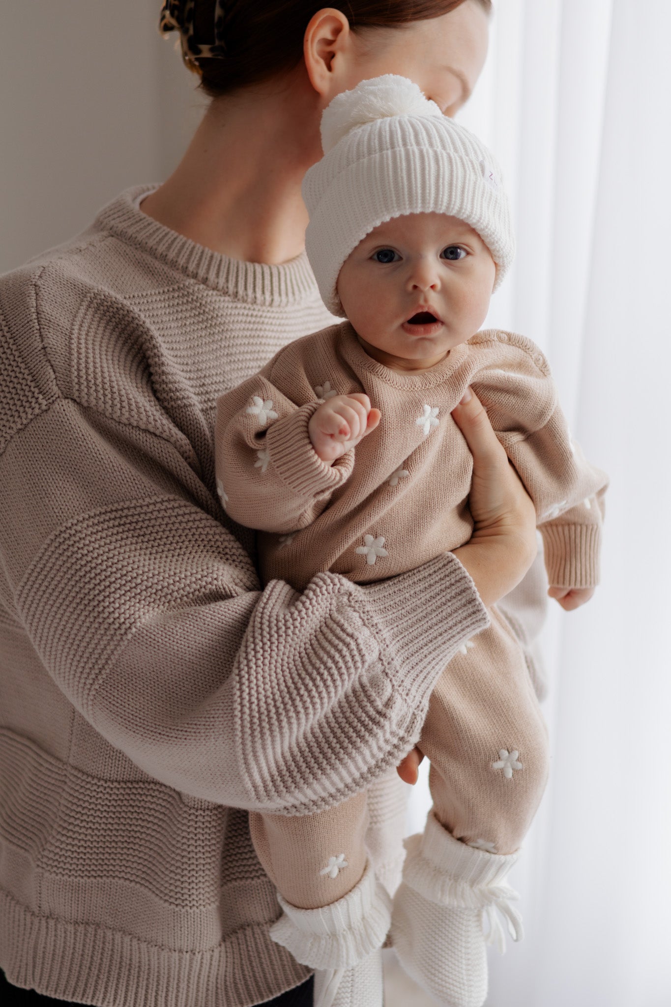 A woman holds a baby in the ZIGGY LOU Ziggy Lou - Winter Playsuit Aurora (Size NB), paired with a white pom-pom hat. The cozy organic cotton playsuit glows in soft natural light as the baby gazes wide-eyed at the camera.