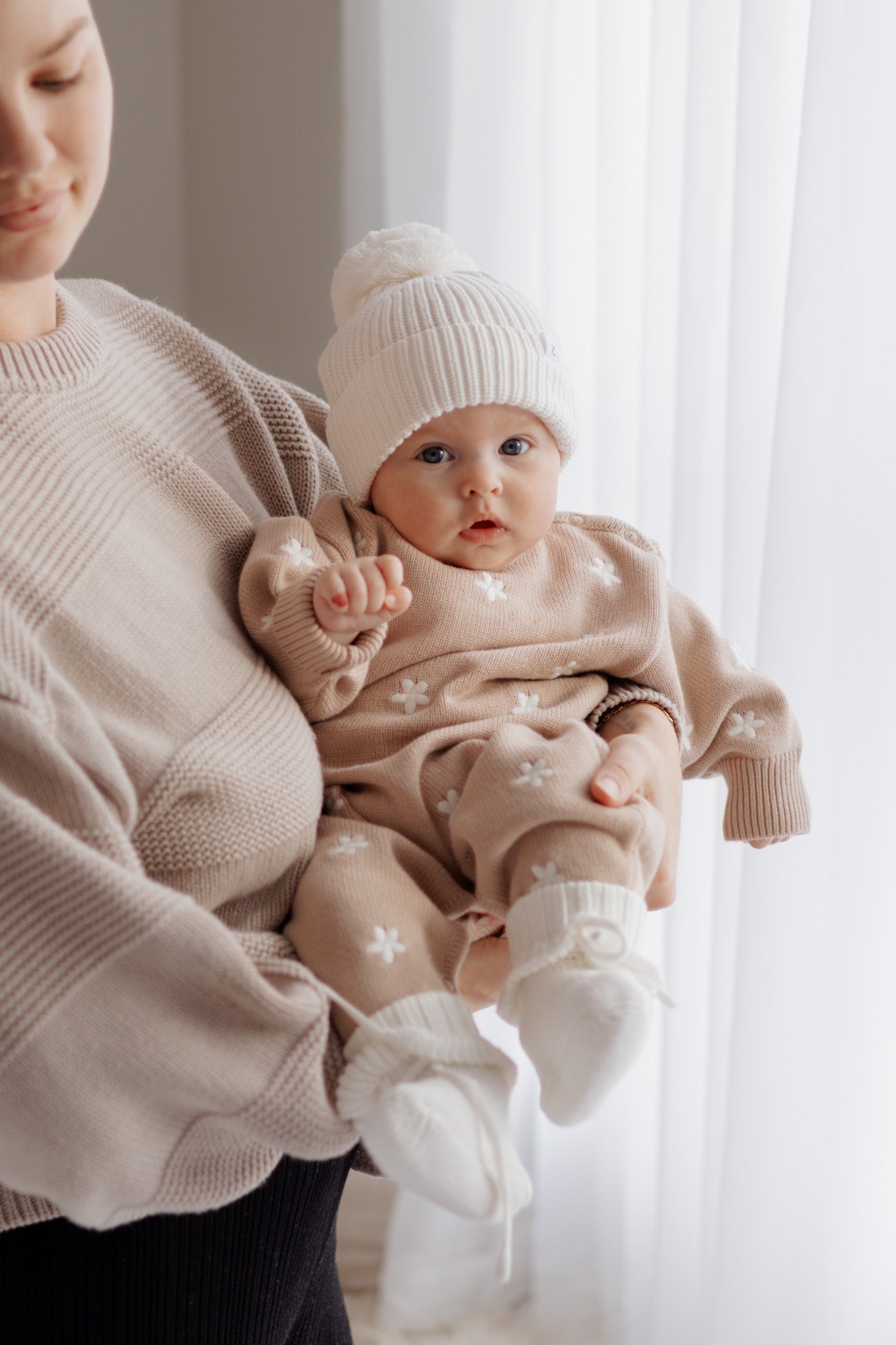 A baby wearing the ZIGGY LOU Winter Playsuit Aurora in size NB, with white booties and a white knit hat, is held by an adult in a beige sweater near white curtains in a gently lit room.
