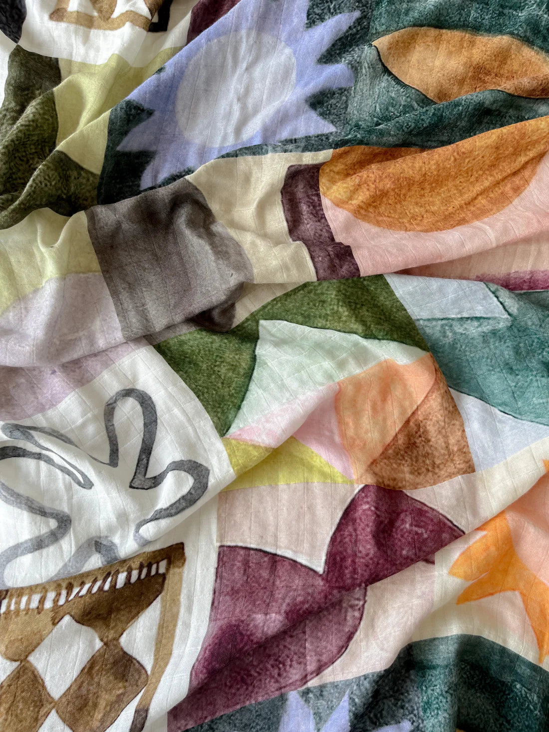 A close-up of the Milky Designs - Sun Checkers Wrap by MILKY DESIGNS, featuring exclusive, hand-painted abstract prints with leafy and geometric shapes in green, orange, purple, brown, and white on slightly wrinkled fabric.