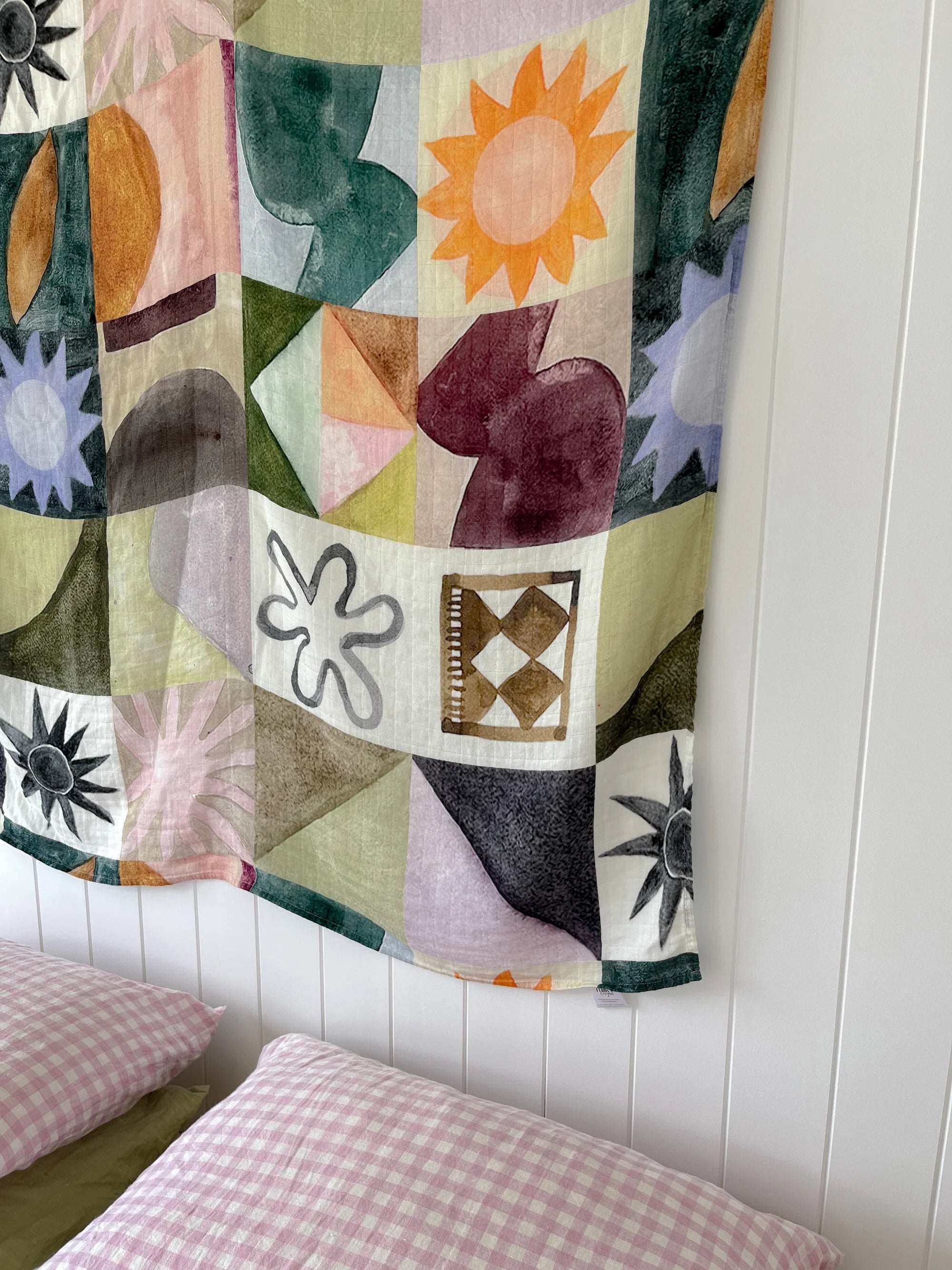 The Milky Designs - Sun Checkers Wrap by MILKY DESIGNS, featuring exclusive abstract prints, sunbursts, and florals, is displayed on a white wall above a bed with two pink and white gingham pillows.