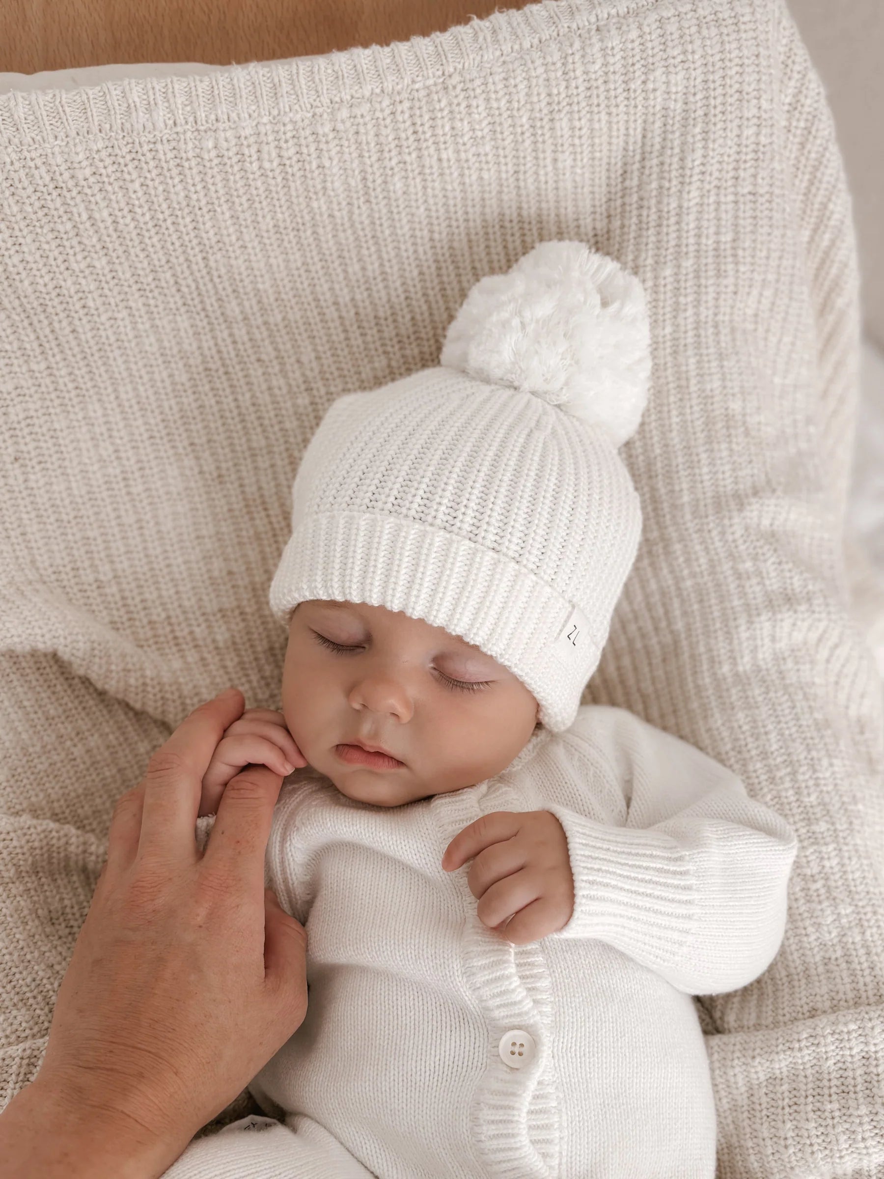 A sleeping baby in a white 100% cotton knit outfit and the ZIGGY LOU Ziggy Lou - Beanie Milk lies on a textured cream blanket, while an adult gently holds the baby’s hand.