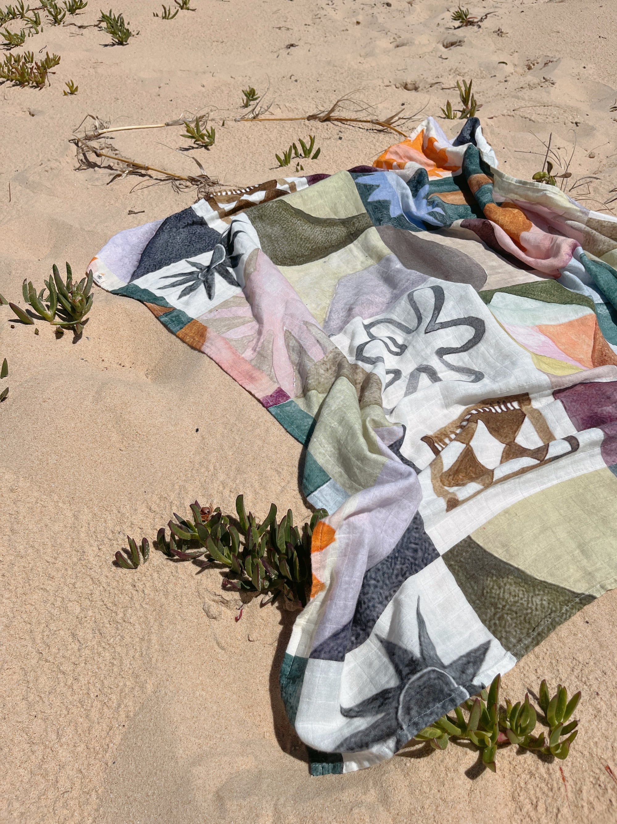 The MILKY DESIGNS Sun Checkers Wrap, a vibrant scarf with exclusive abstract prints, rests on sandy beach ground, partially covering green plants and surrounded by dry sticks.