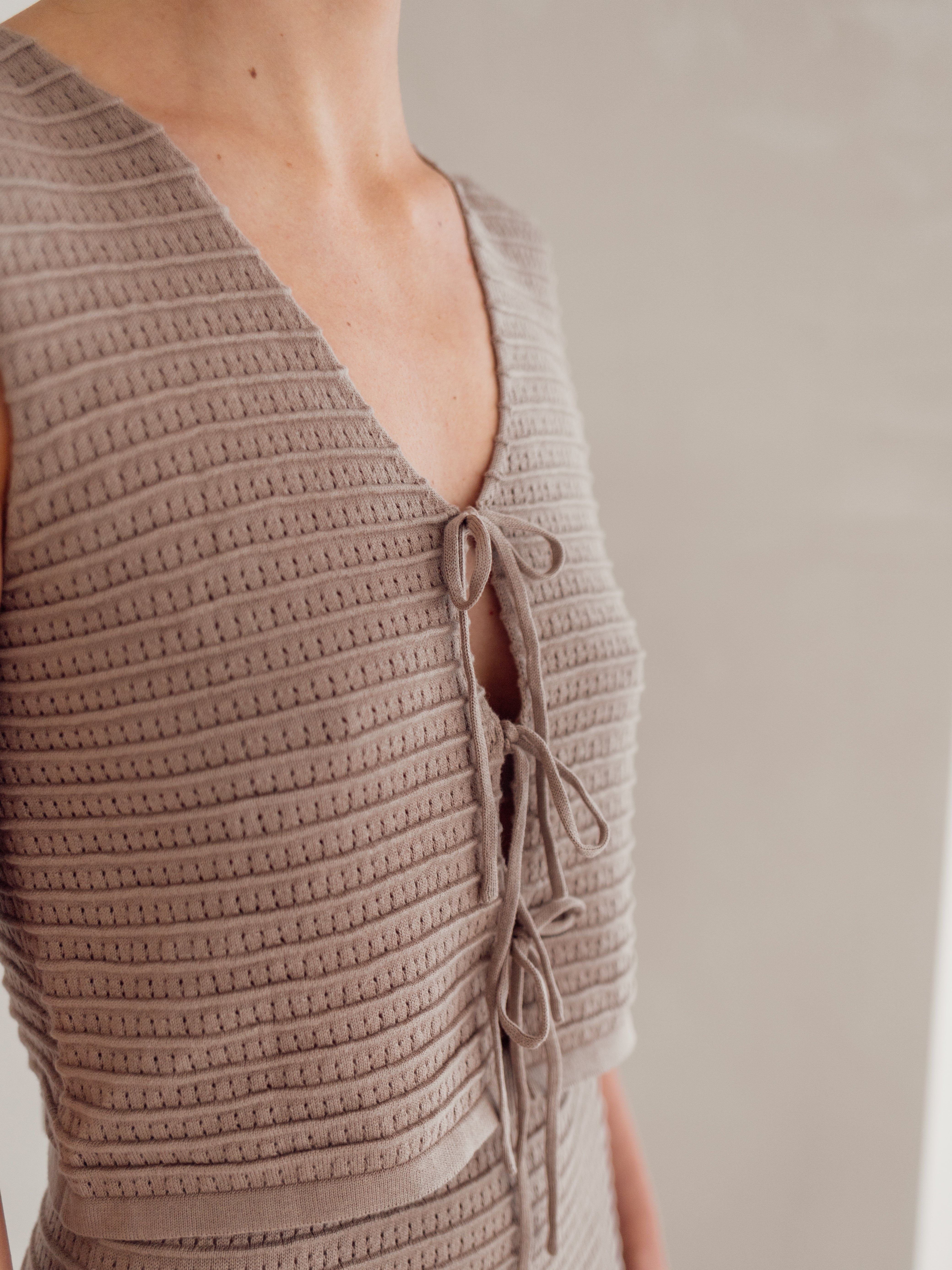 A person is wearing the Women's Knit Singlet Milo, a textured, sleeveless beige top from ZIGGY LOU, made from organic cotton. It features a V-neck and lace-up detailing in the front, set against a neutral, light-colored background.