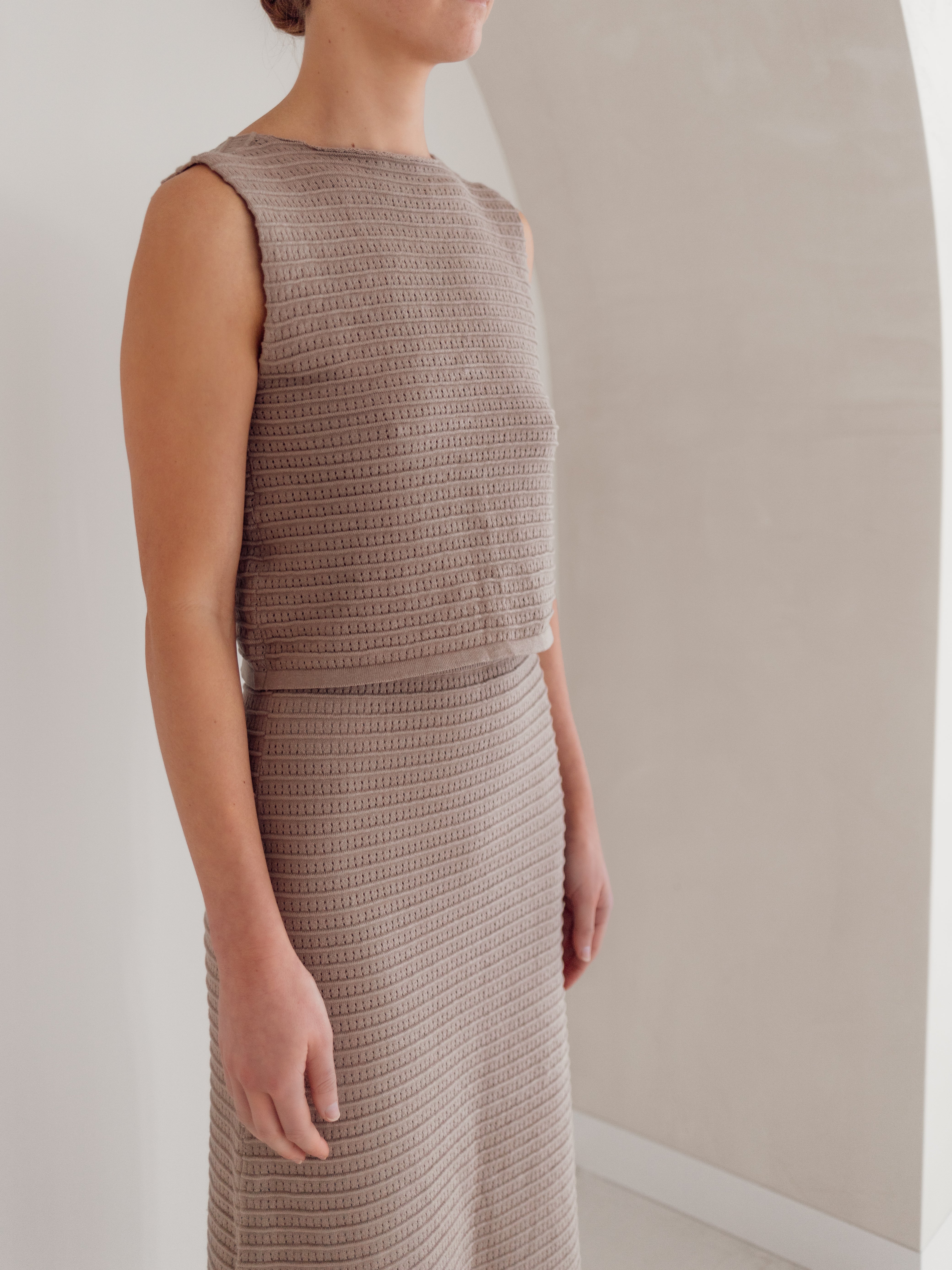 A person wearing the Women's Knit Singlet Milo by ZIGGY LOU stands in a minimalist setting with a white arch in the background. The focus is on the singlet's textured pattern and adjustable ties, highlighting the person's elegant posture.