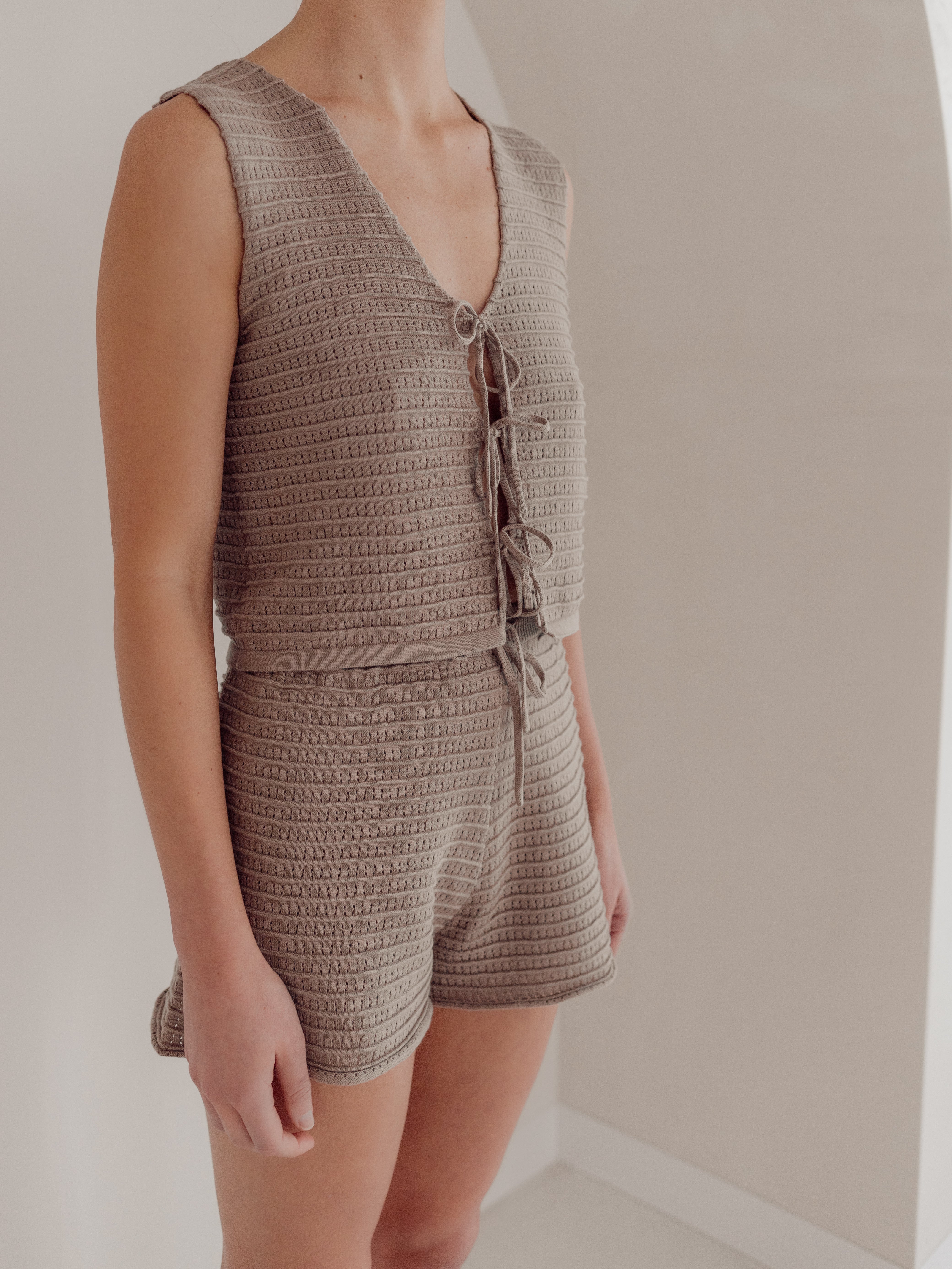 A person is wearing ZIGGY LOU's Milo Women's Knit Shorts and a matching sleeveless top. The beige, 100% organic cotton top features a V-neck with tie-up details. They are in a light-colored, minimalist interior with an archway.