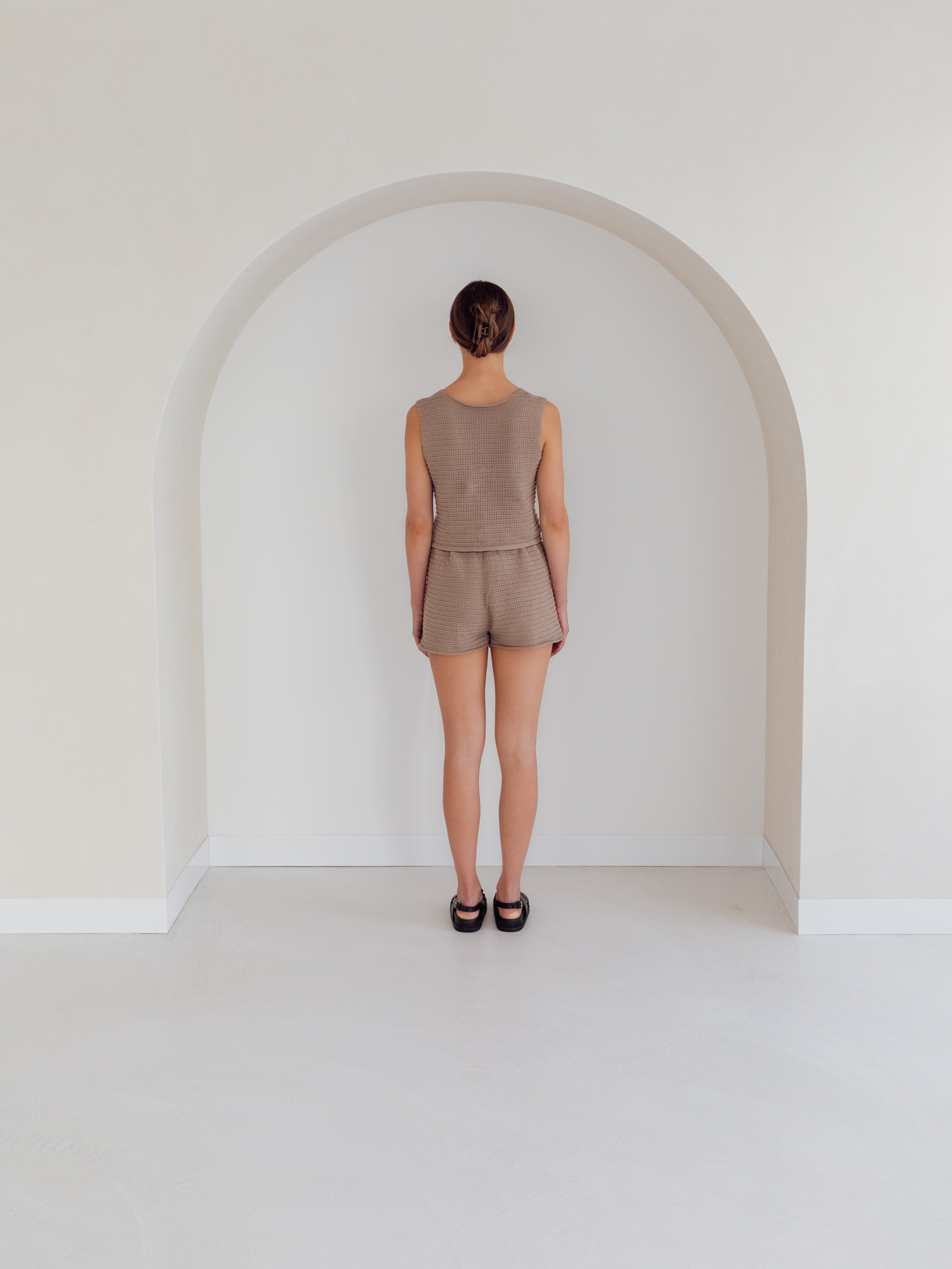 A person with brown hair stands facing a white wall inside a small, arched alcove. They are wearing the Women's Knit Singlet Milo and matching shorts from ZIGGY LOU, along with black shoes. The minimalist setting enhances the natural elegance of their attire.