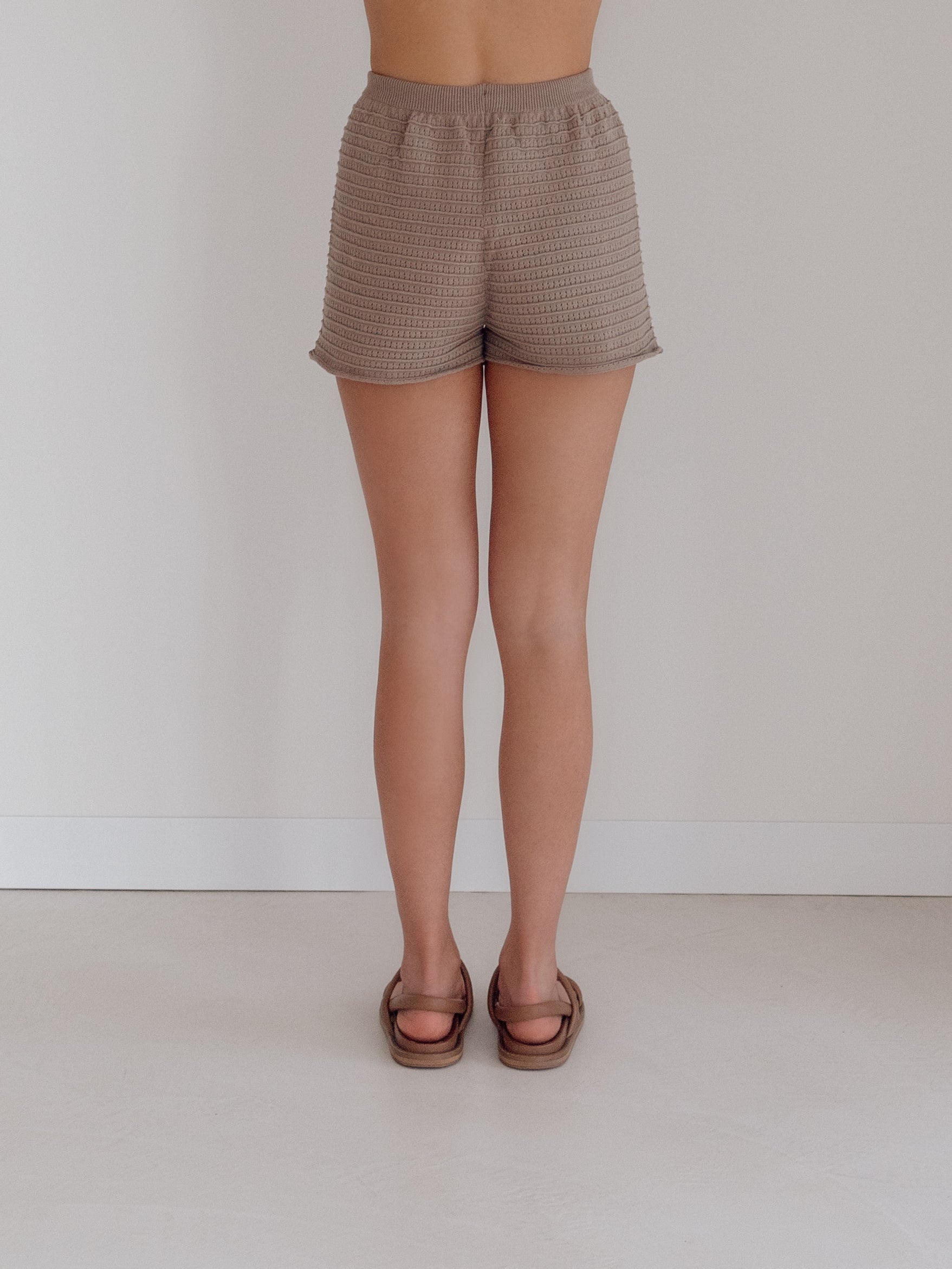 A person stands facing a white wall in textured beige Women's Knit Shorts Milo by ZIGGY LOU and brown sandals. The image captures mid-back to feet on a light floor, showcasing the comfortable elegance of 100% Organic Cotton.