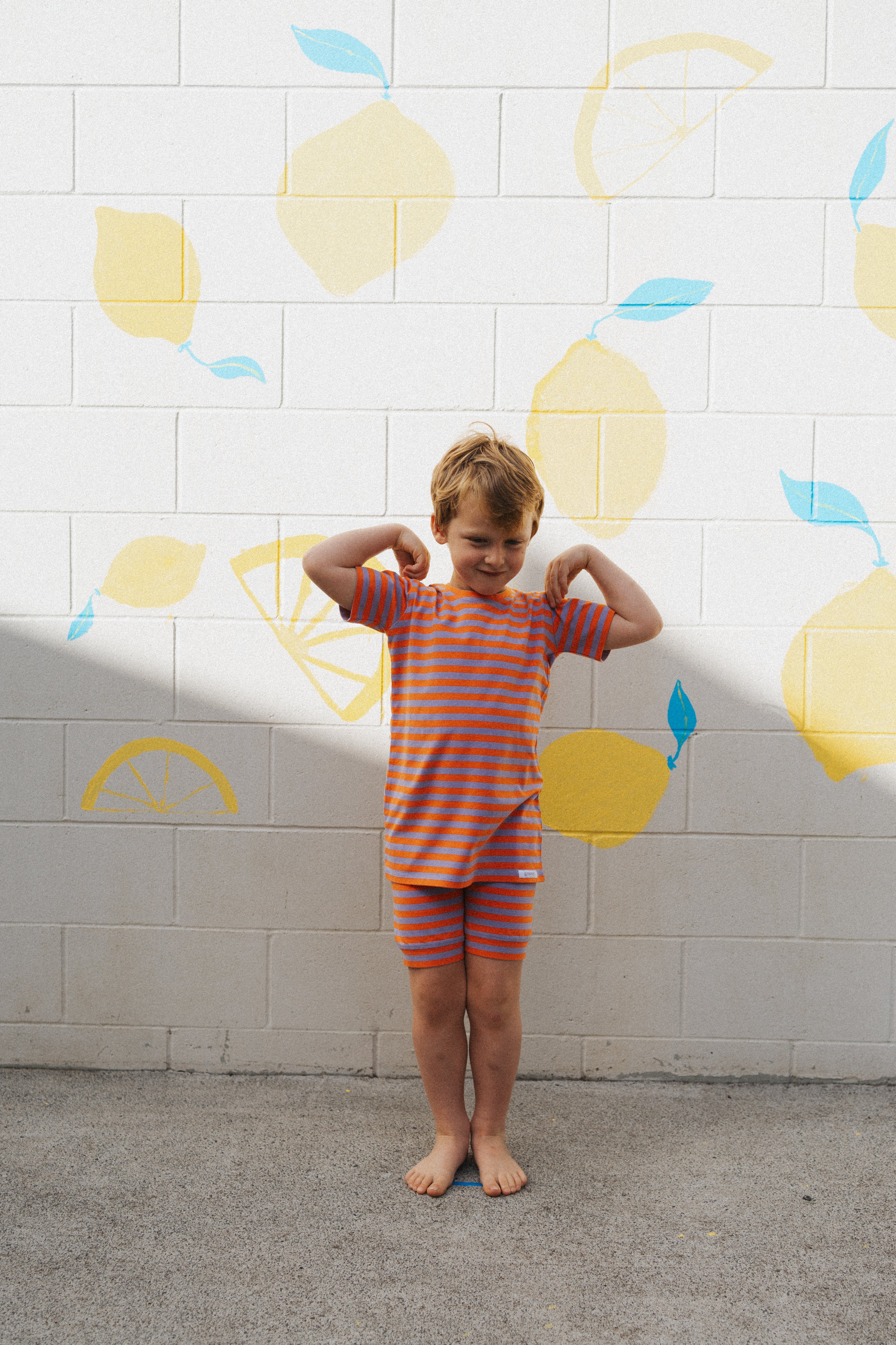 A young child wearing the G.NANCY Stripe Shortie PJ Set in Jacaranda/Peel playfully flexes their arms in front of a white brick wall adorned with pastel yellow lemons and blue leaves. The snug-fitting outfit, made from organic cotton jersey, is ideal for barefoot fun on the cool concrete surface.