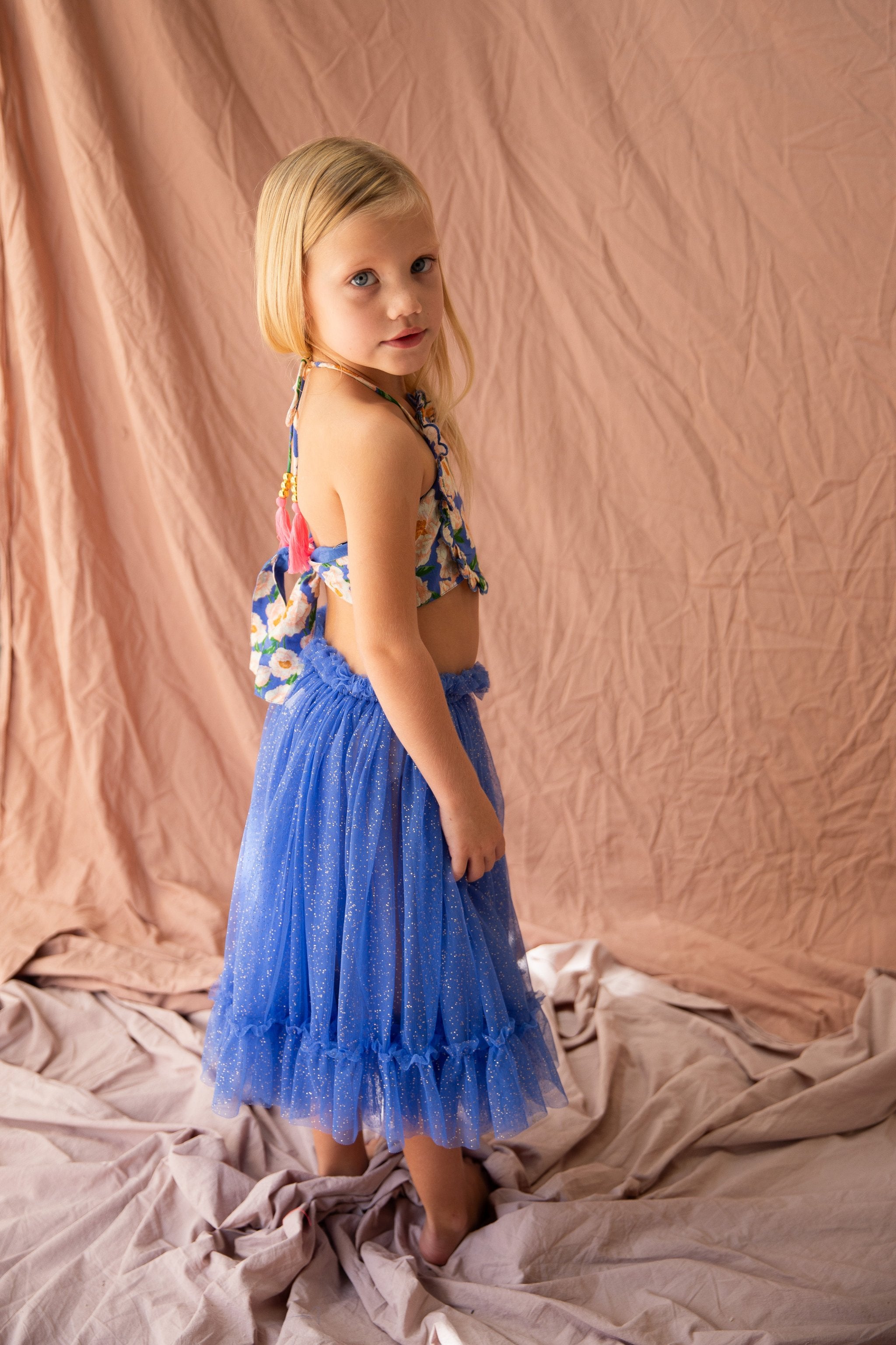 A young girl with blonde hair is dressed in the Jess Top Soirée Bouquet/Bondi Blue by BELLA + LACE, paired with a blue sparkly skirt. She stands against a light brown, wrinkled fabric backdrop, looking over her shoulder towards the camera with a neutral expression.