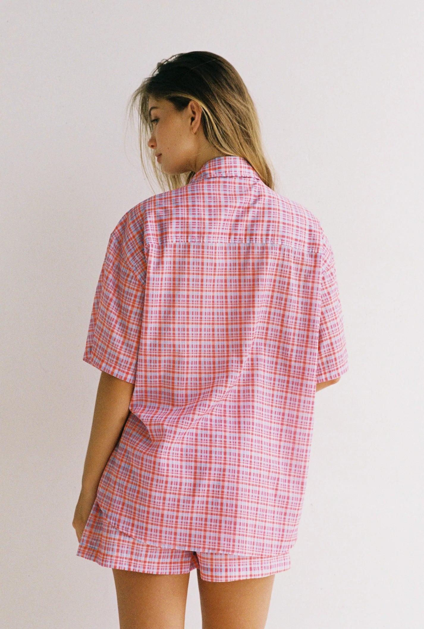 A woman with long hair stands facing away, wearing the JUNI Watson Shirt—an oversized, short-sleeved pink and red plaid button-up by JUNI—paired with matching shorts against a plain, light background.