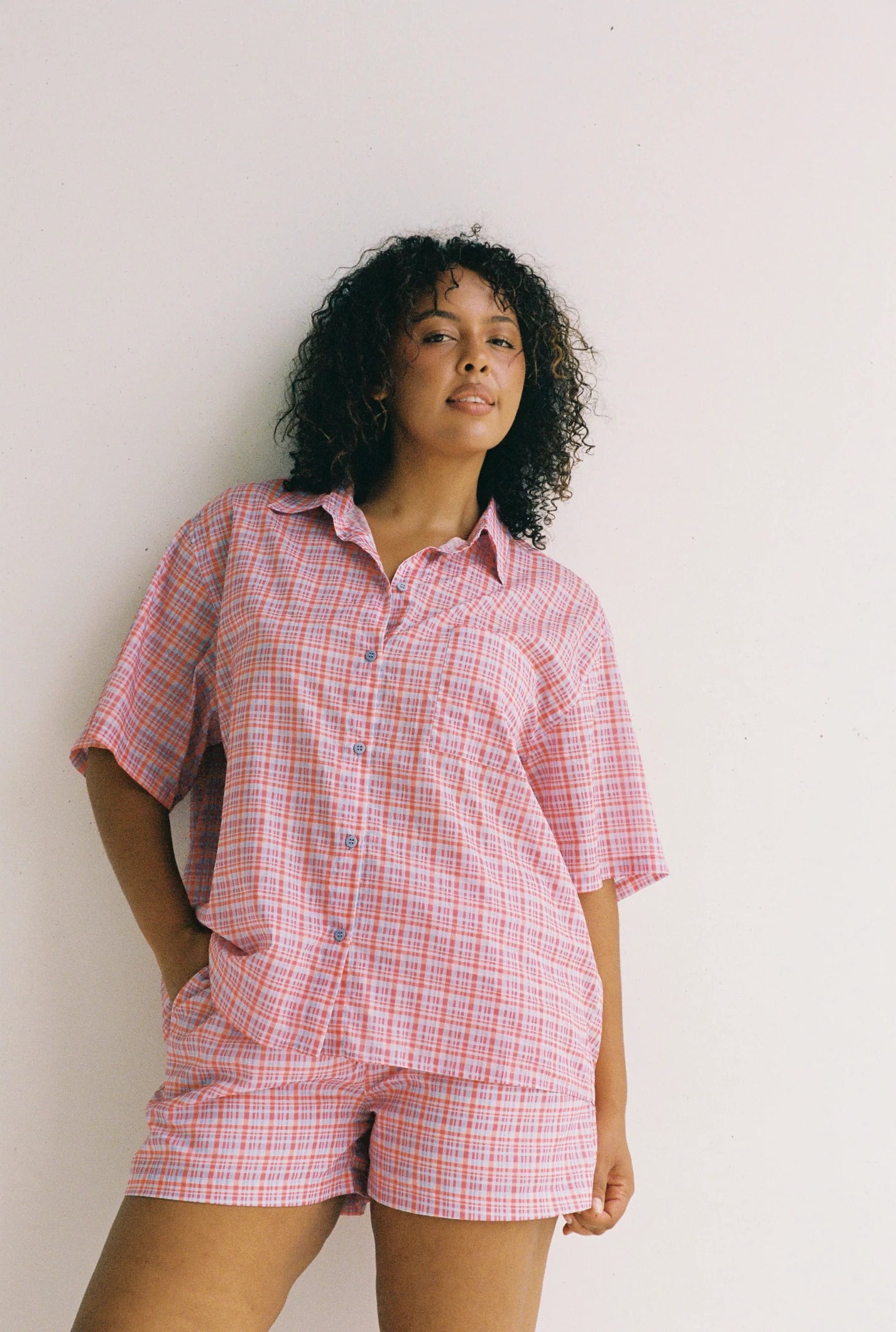 A person with curly hair wears the ethically made JUNI Watson Shirt in pink and white plaid, standing against a plain white background with one hand in their pocket.