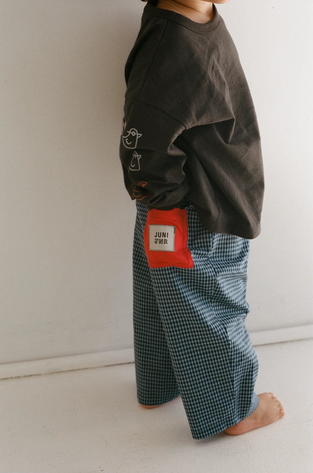 A child stands barefoot against a white wall, wearing a dark long-sleeve shirt and JUNI JNR Benji Pant Blue Check, 100% cotton pants with an elastic waistband and red patch on the pocket. The child's face is not visible.