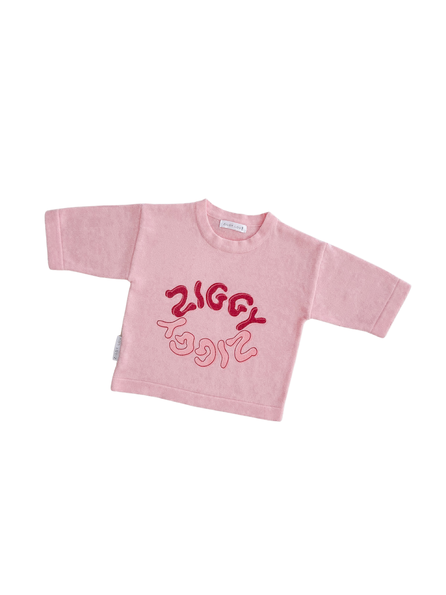 The Ziggy Lou Long Sleeve Tee Thea for toddlers is light pink, features bold wavy red "ZIGGY" text and abstract designs, is made from 100% cotton, and is shown on a plain black background. Brand: ZIGGY LOU.