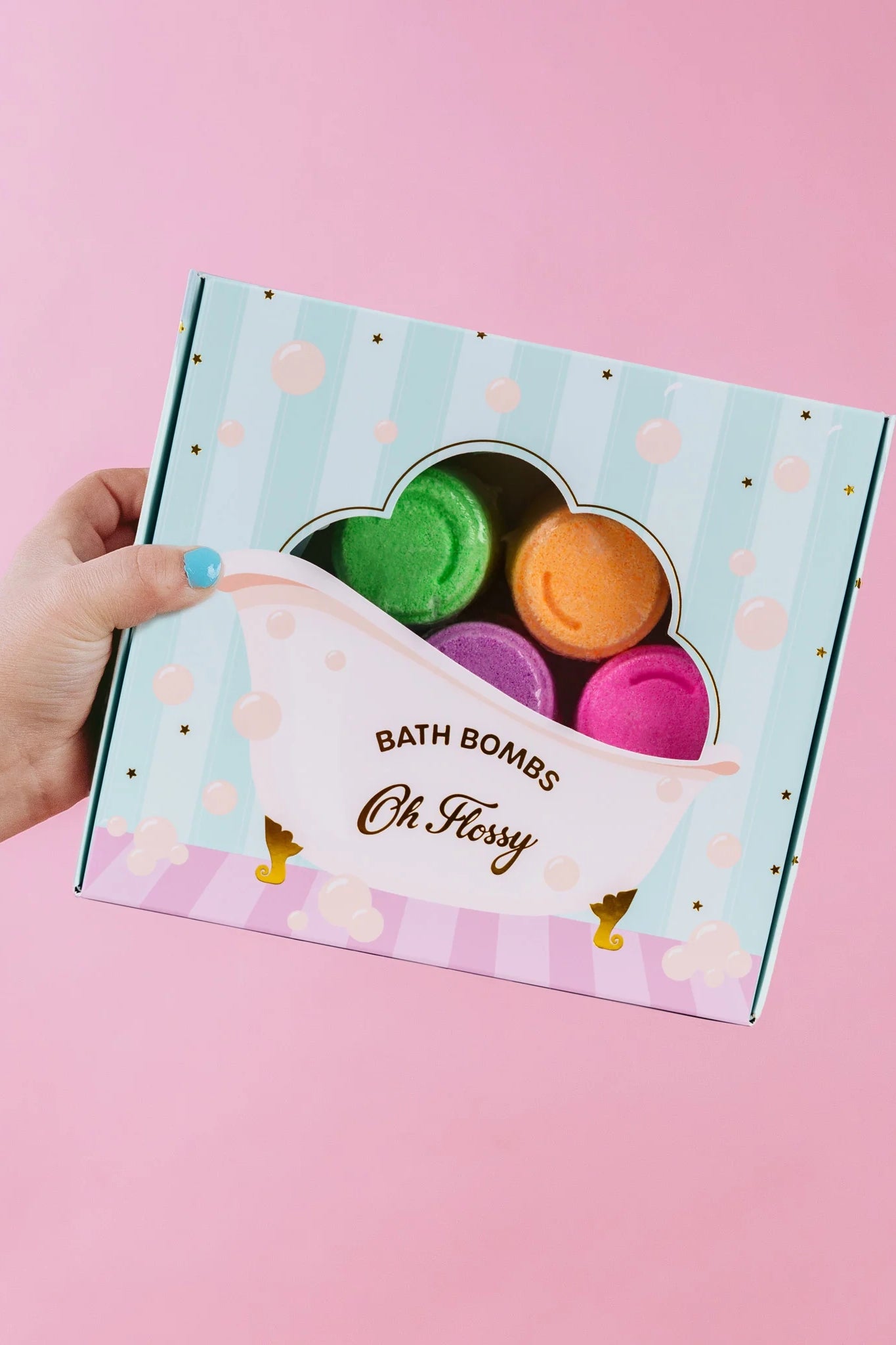 A hand with blue nails holds an Oh Flossy Kids Mini Bath Bombs box, styled like a clawfoot tub, against a pink backdrop. Inside are green, purple, orange, and pink bath bombs. Handmade in Australia by OH FLOSSY, they are gentle on kids' skin.