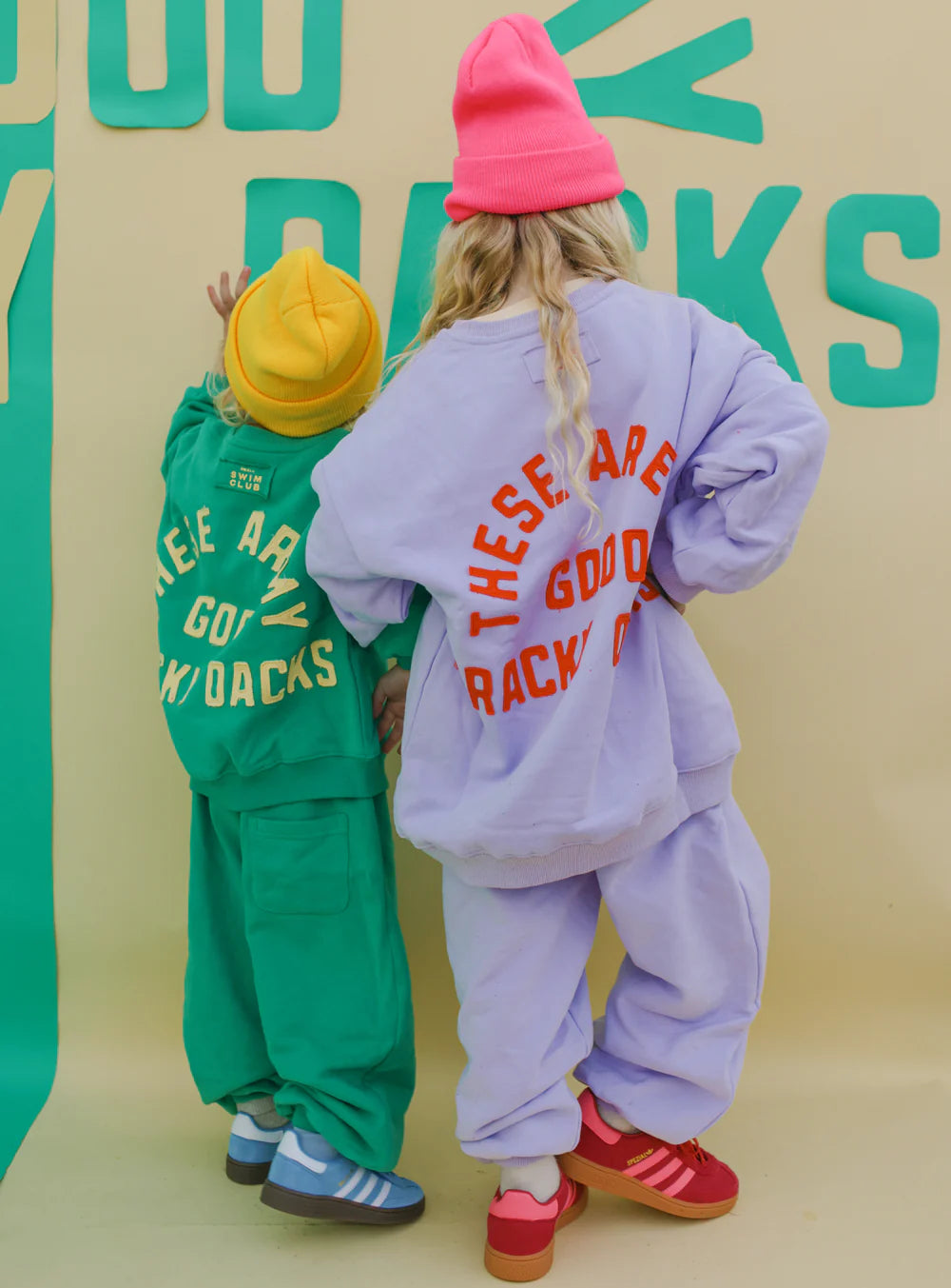 Two kids face away from the camera, wearing SMALL SWIM CLUB Good Tracky Pullovers in lavender/red and beanies. Their clothes read “THESE ARE GOOD SNACK DAYS,” set against a bold background with lively green and yellow text graphics.