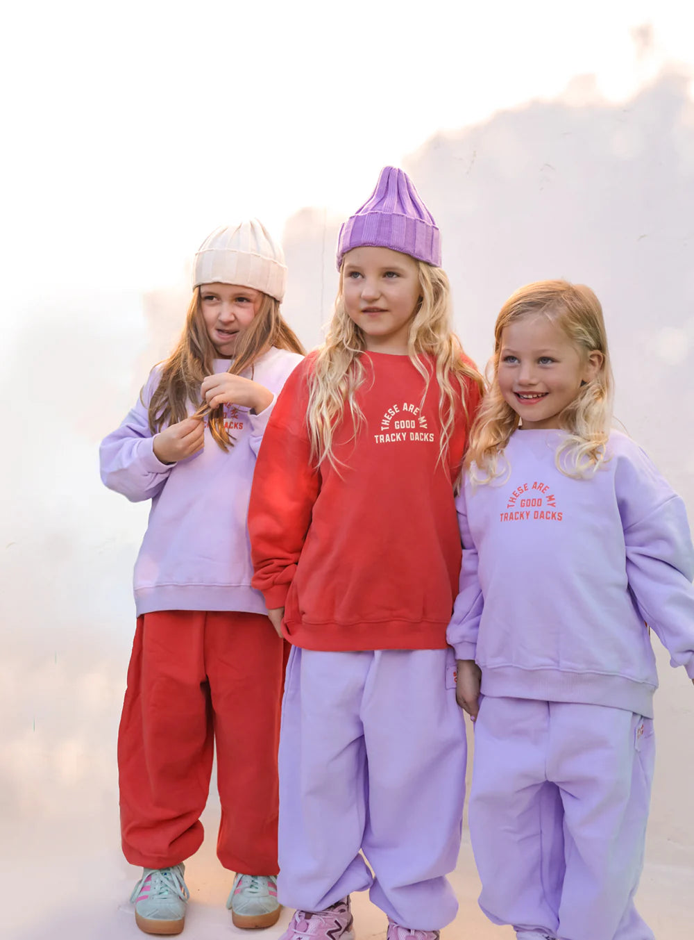 Three girls wear SMALL SWIM CLUB’s Good Tracky Pullover in Lavender/Red with matching trackies. Two have lavender sets, one wears a red pullover; all have long blonde hair, and one tops off the look with a knit hat.