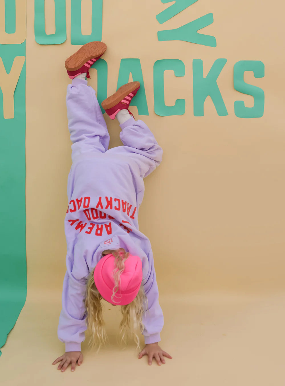 A young child in a pink beanie and SMALL SWIM CLUB Good Tracky Pullover in Lavender/Red does a handstand against a beige wall with teal and yellow letters, legs bent and red shoes resting for support.