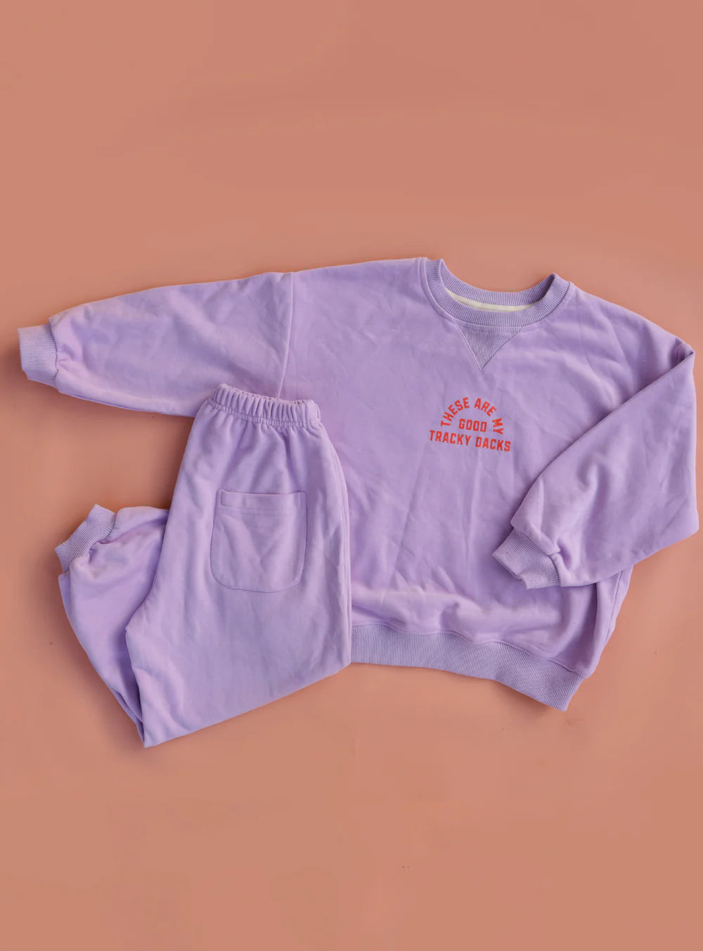 The SMALL SWIM CLUB Small Swim Club - Good Tracky Pullover Lavender/Red is a soft, fleece-lined, slouchy sweatshirt in lavender, shown flat on a peach background with red and yellow “Loose Ace Good Tracky Dacks” text on the front.
