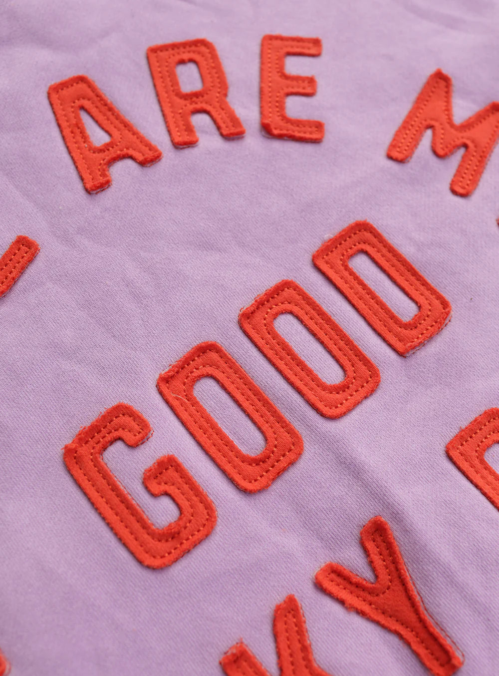 Close-up of red stitched "GOOD" letters on lavender fabric, from the SMALL SWIM CLUB Small Swim Club - Good Tracky Pullover. Features playful textures and soft fleece lining for added comfort in this cozy design.